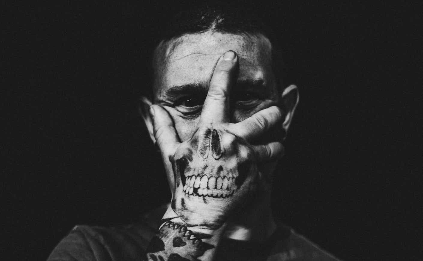 A man covers his face with his hand, which has a skull tattoo