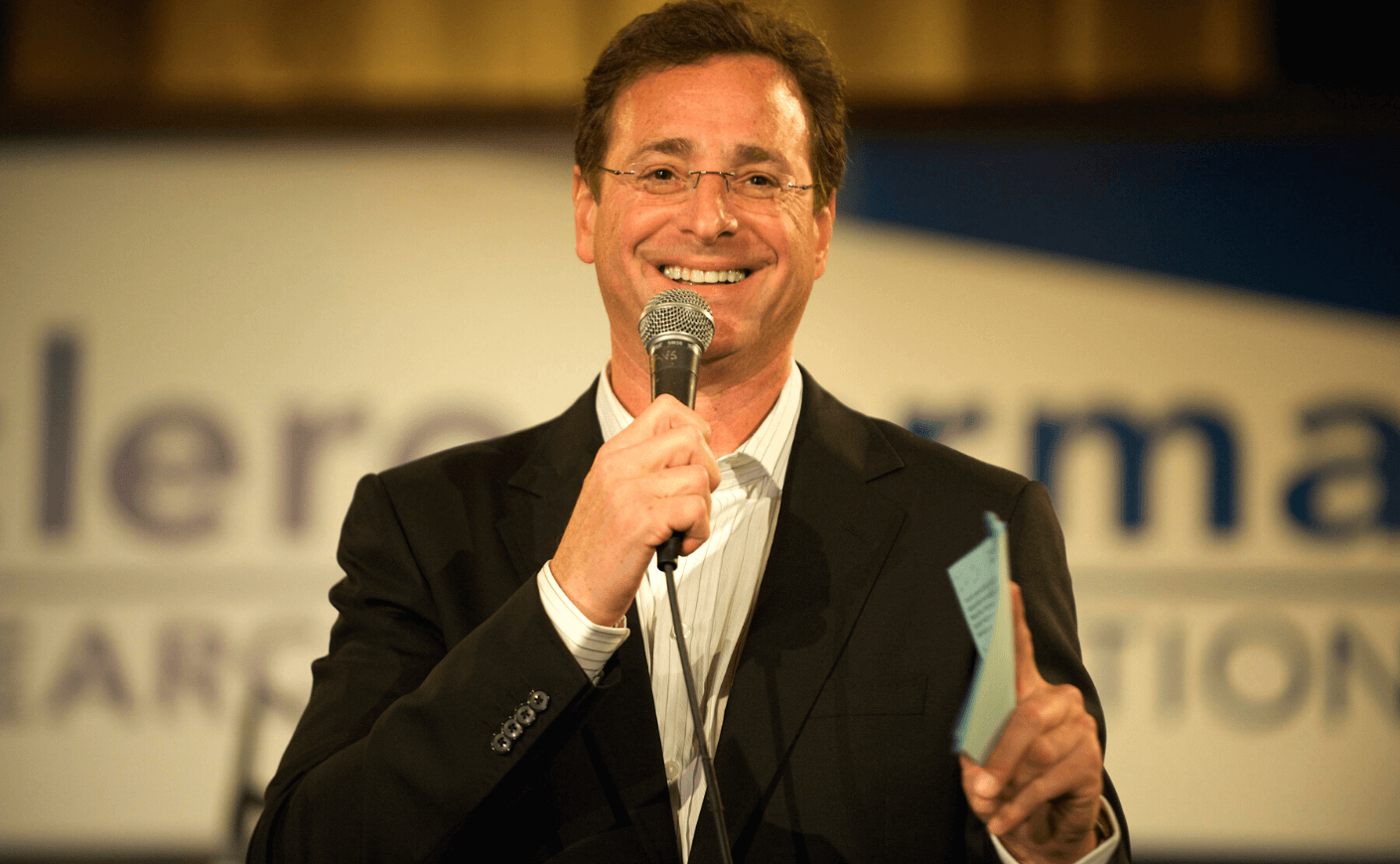 Comedian Bob Saget holding a microphone and card