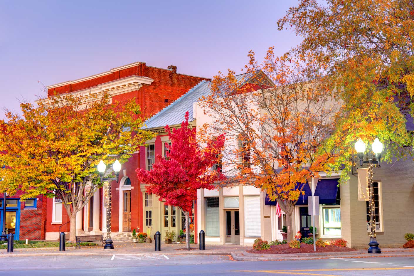 Leafy, red autumn trees pictured in front of quaint, old-fashioned downtown, one-story buildings.