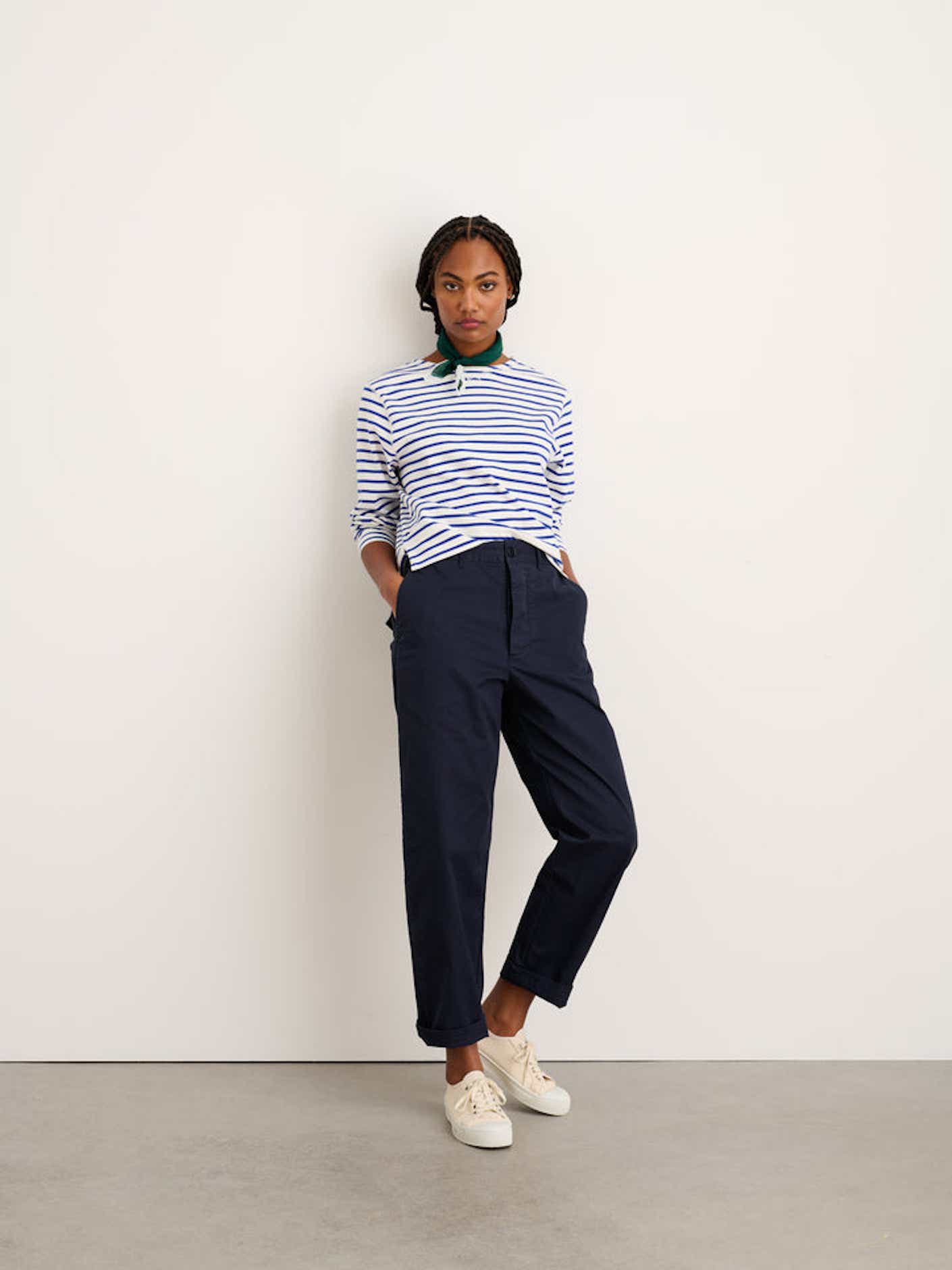 A woman wearing navy blue chinos leans against a white wall.