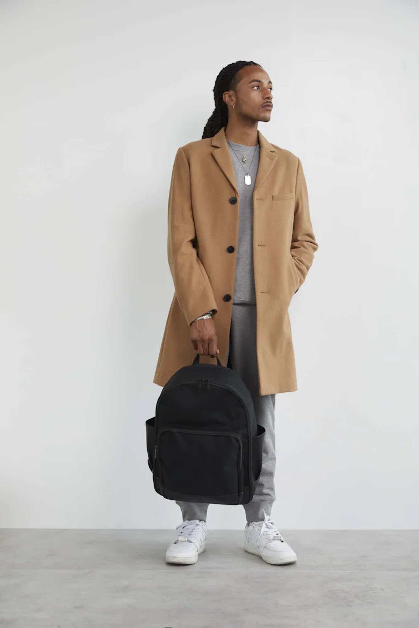 A person wearing a trench coat is holding a minimalistic black backpack.