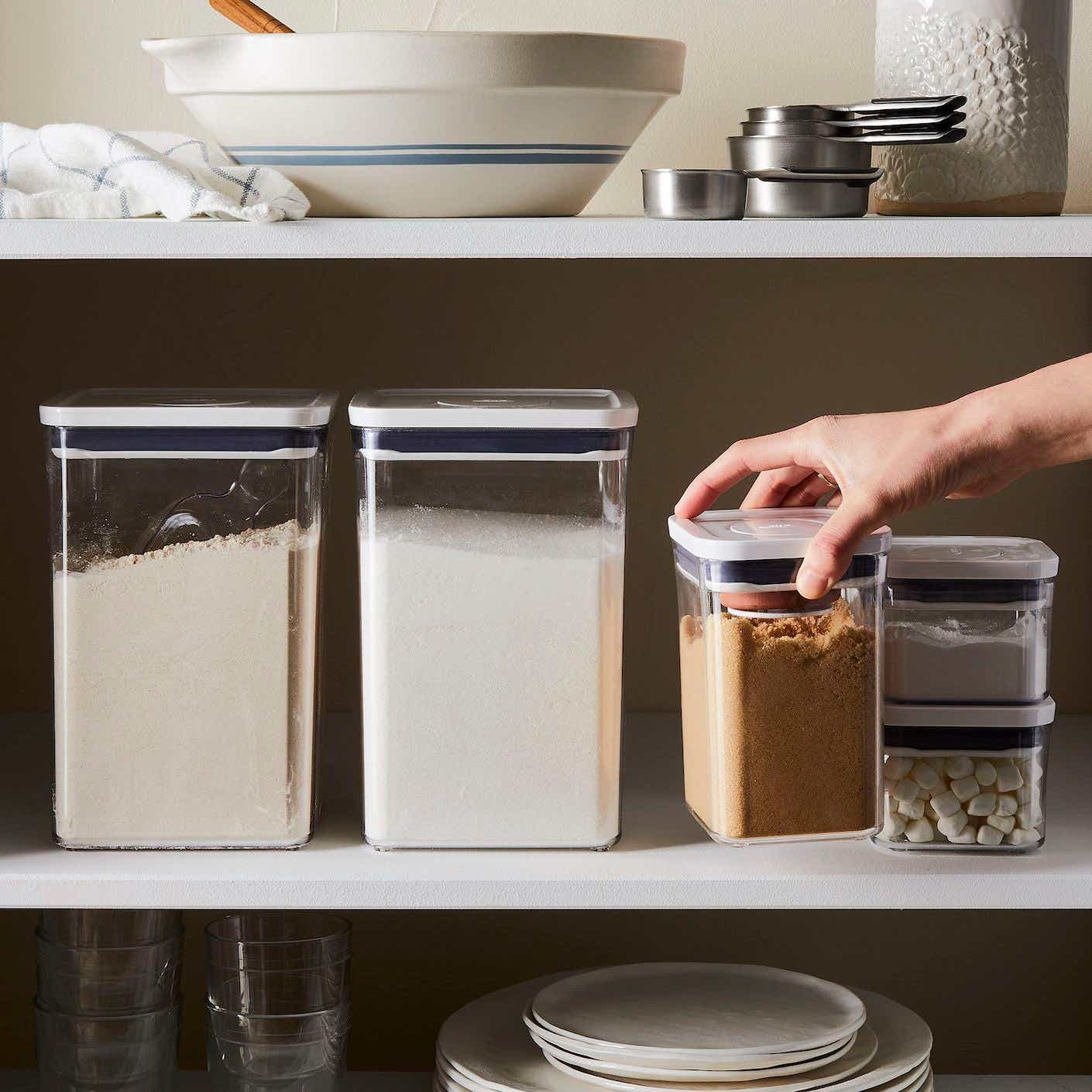 A hand pulls a clear food storage container off of a full pantry shelf.