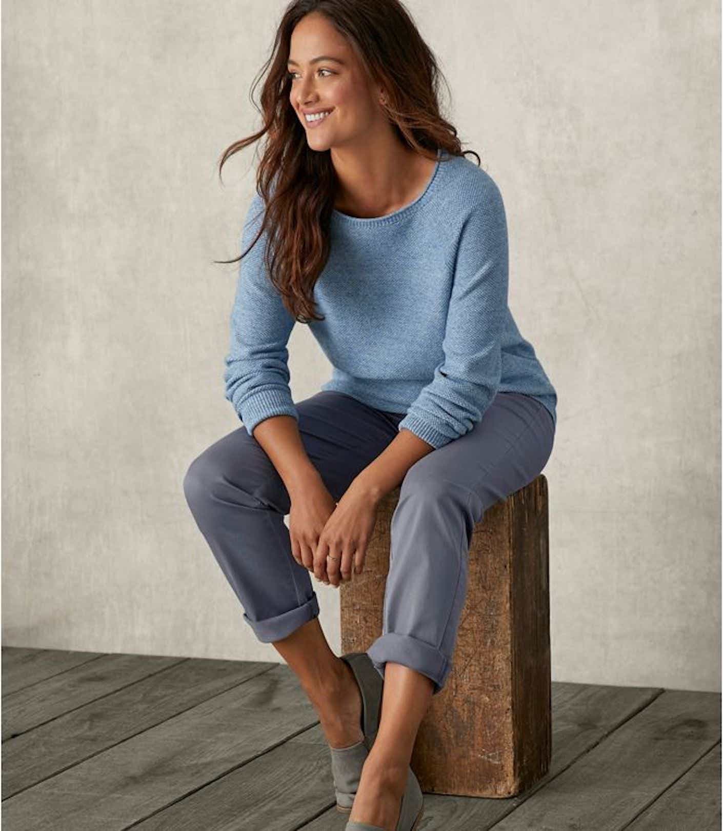A smiling woman wearing blue chinos sits on a block of wood.