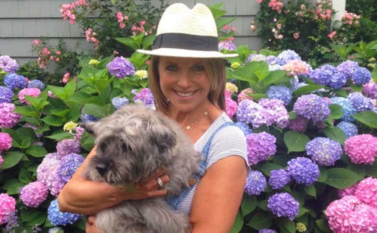 Katie Couric stands in front of a hydrangea shrub holding her dog and wearing a straw hat with a black ribbon around it