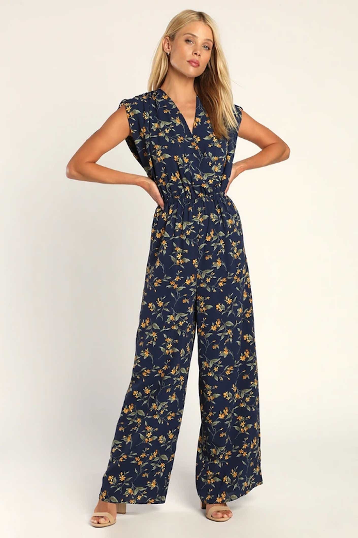 A woman poses with her hands on her hips in a long, flowy, flutter sleeved jumpsuit that is black and covered in a small floral pattern.
