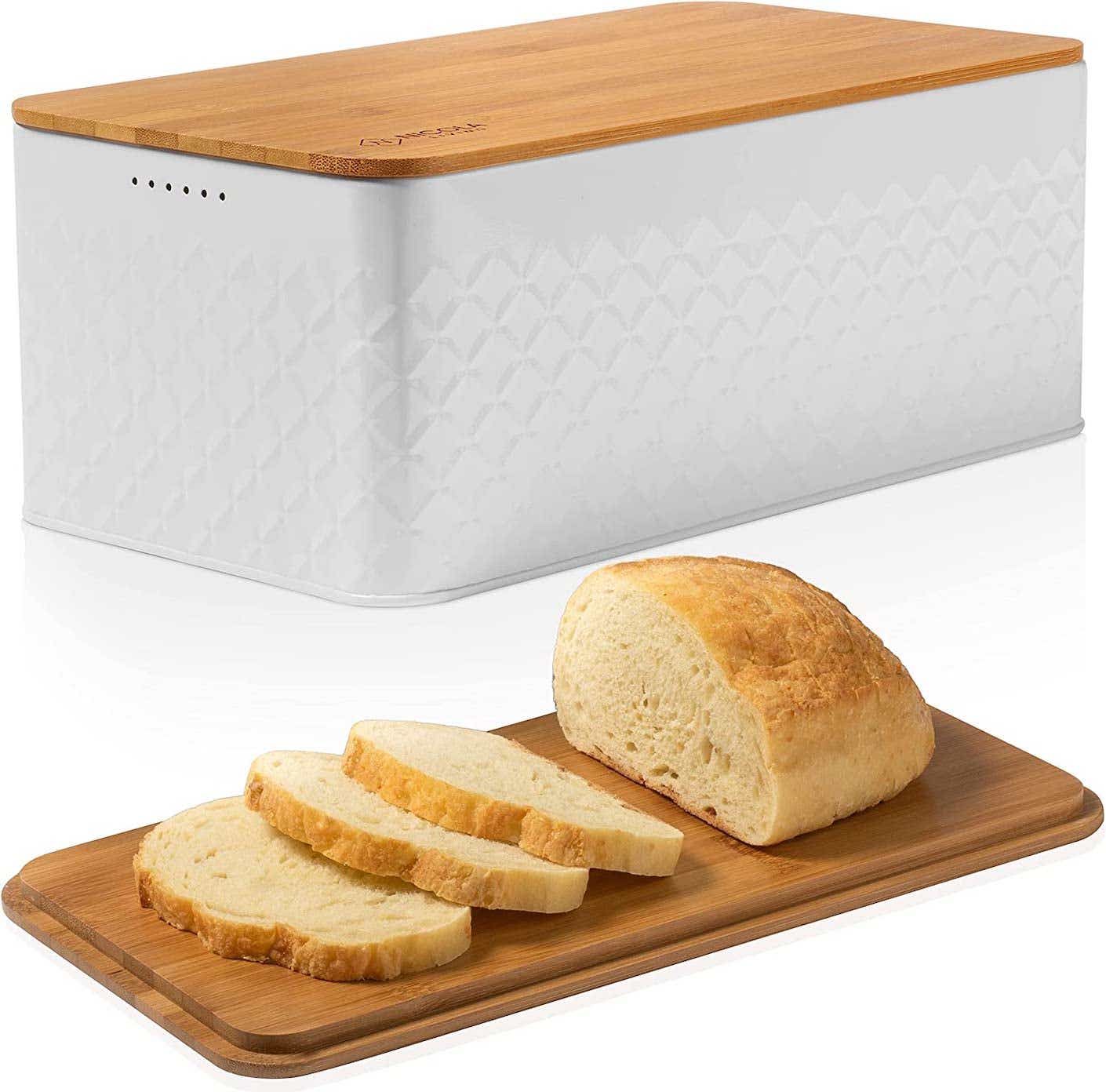 A loaf of bread rests on the wooden cutting board that doubles as a lid for a white metal bread box that sits in the background.
