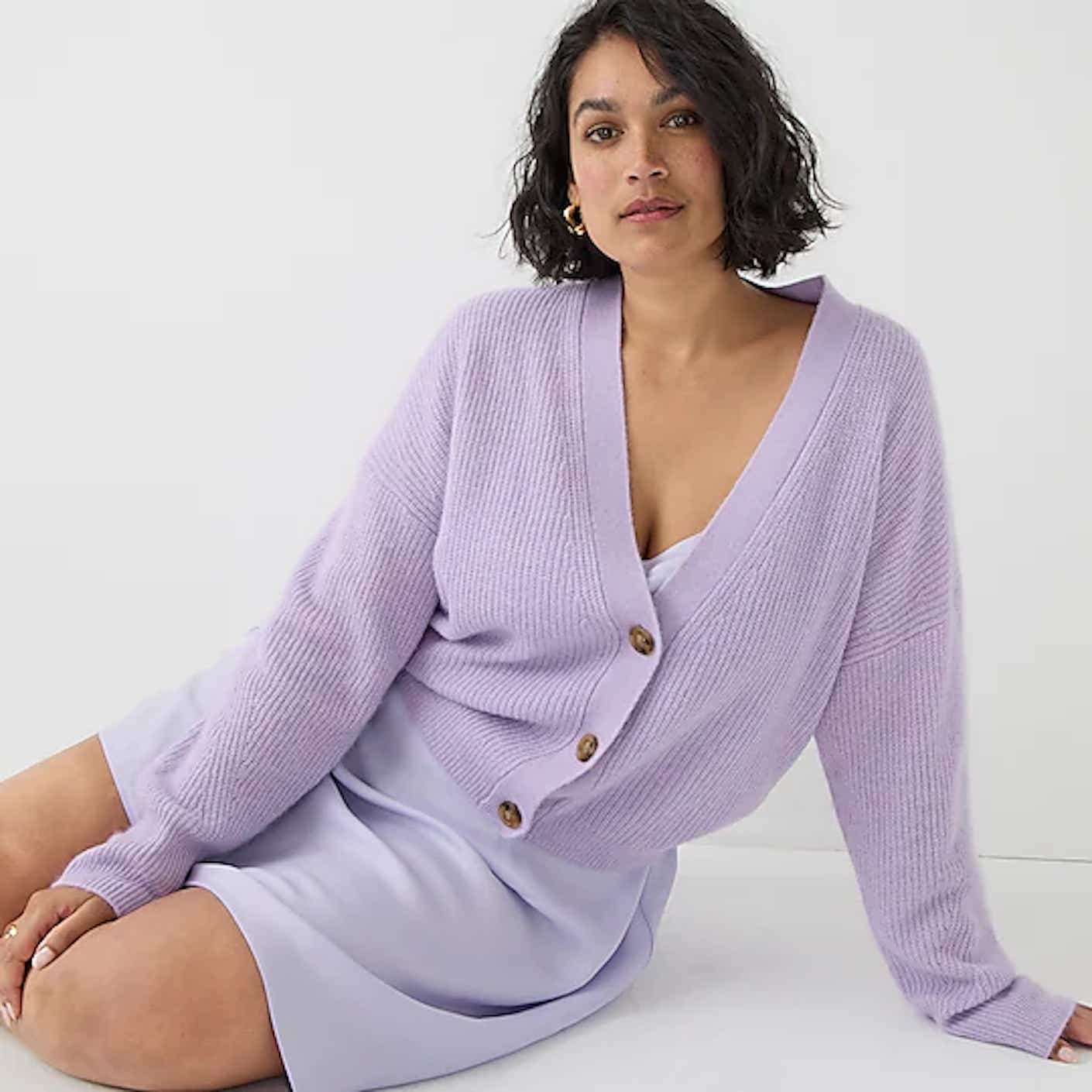 A woman sits in a half-lying down position while wearing a buttoned up, deep V, soft, cozy, light purple cardigan.