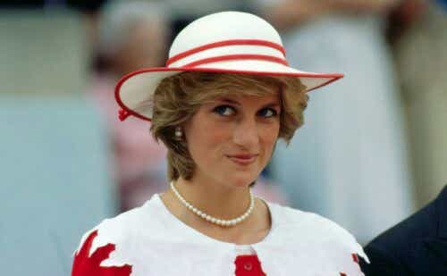 Princess Diana wears a white hat and matching suit with red accents while smiling slyly.