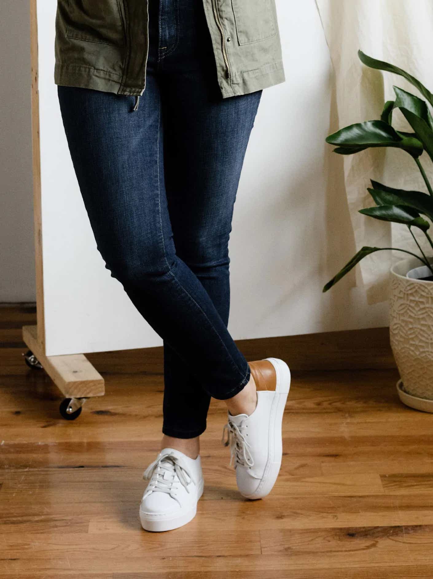 A person wears a pair of white sneakers with leather accents on a hardwood floor, next to a green plant.