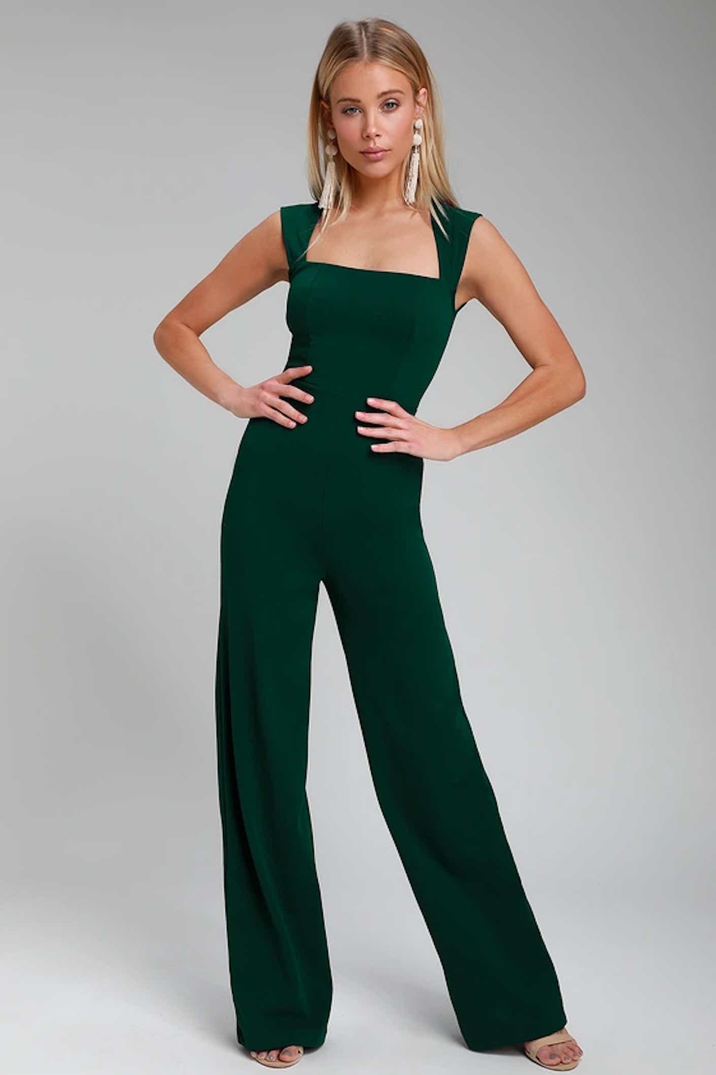 A woman poses with her hands on her hips in a emerald green tank jumpsuit with a square neck.