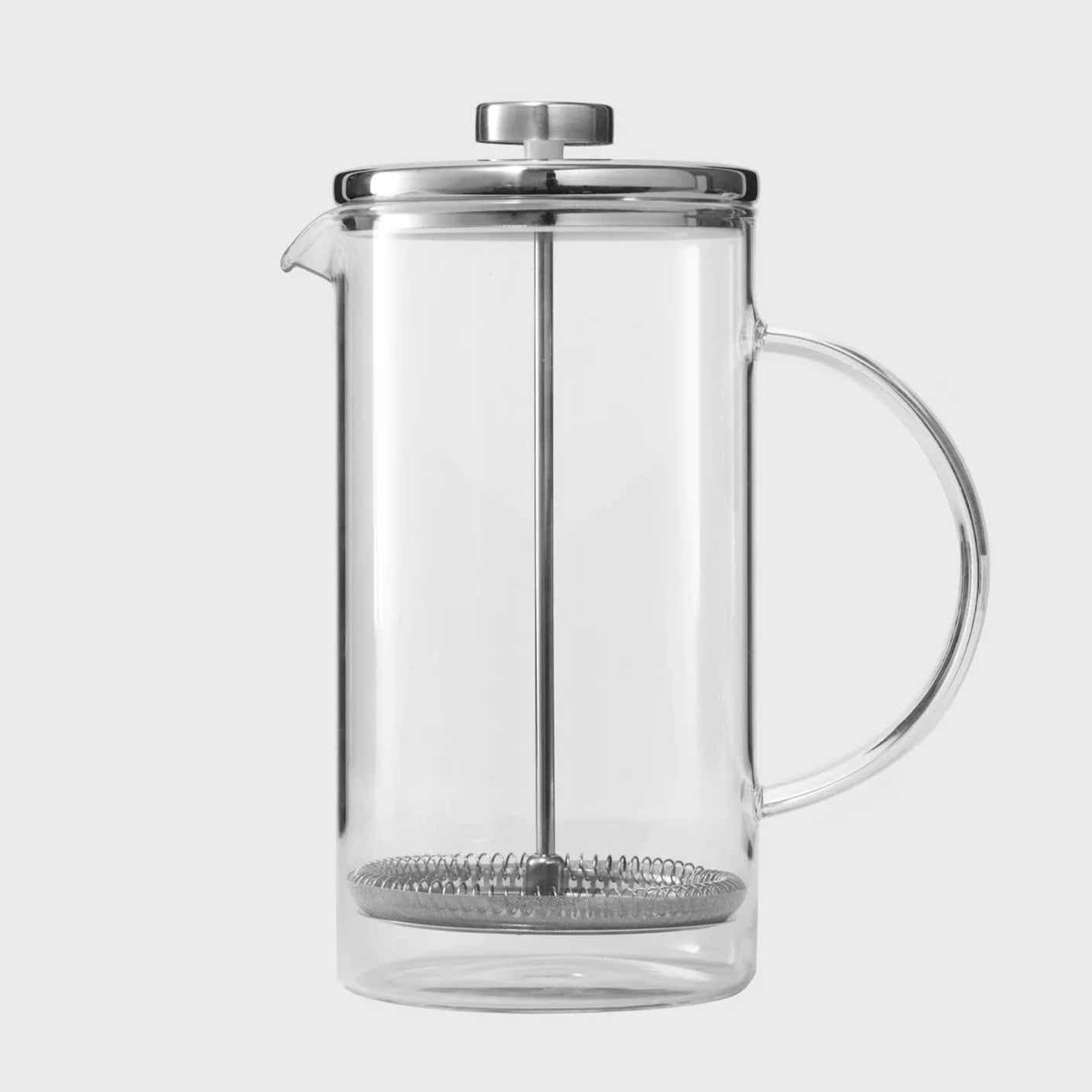 A clear glass French press with a clear handle and a metal lid and strainer is pictured in front of a white background.