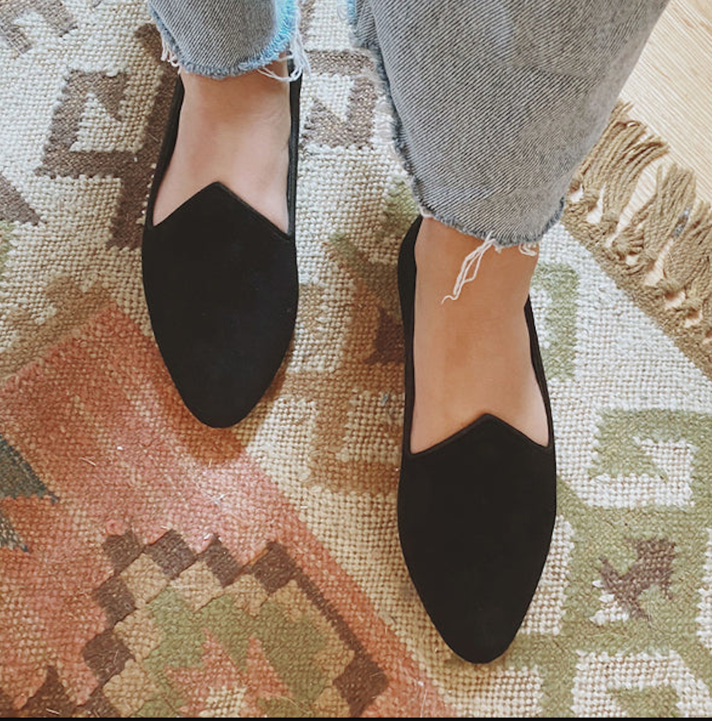 A person wears a pair of black, suede flats with a pair of light blue jeans while stepping on an orange patterned rugs.