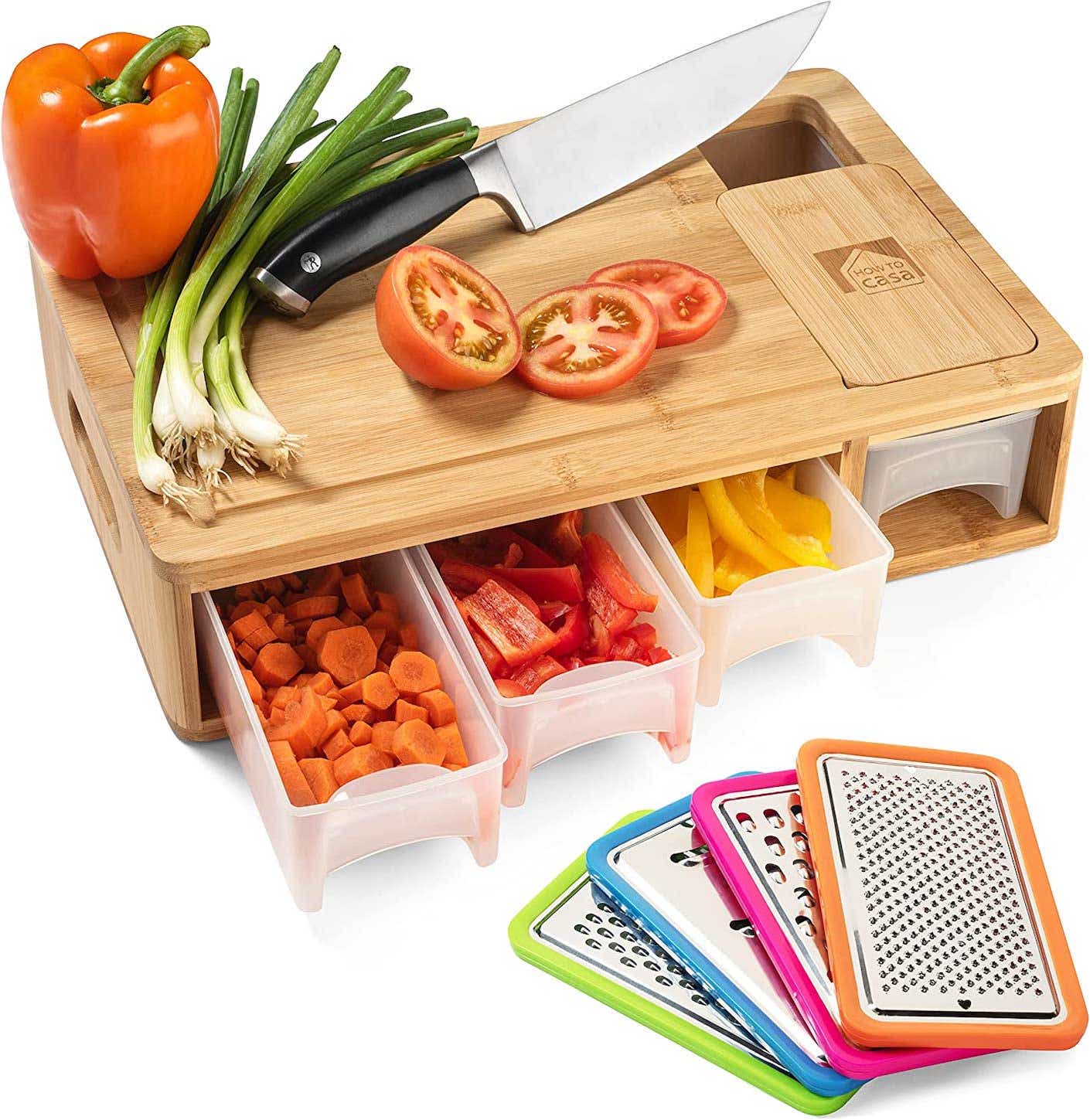 A wooden prep station has a chopping board on top that's strewn with vegetables and four clear plastic compartments below that hold chopped vegetables alongside four grater attachments.