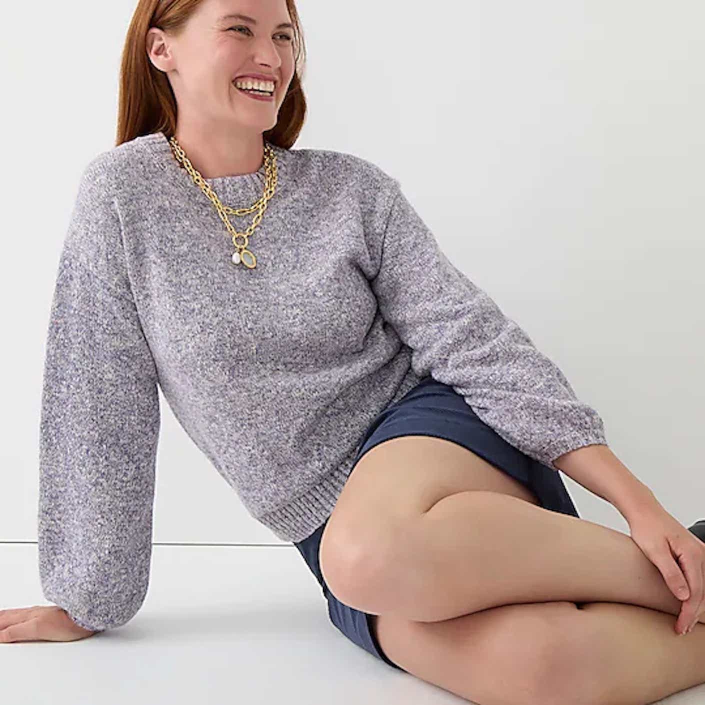 A woman half-sitting up with her knees curled towards the camera wears a light gray, crewneck pullover sweater with slightly puffed long sleeves.