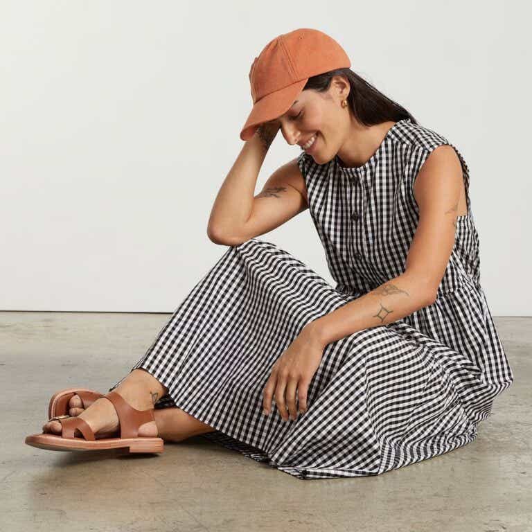 A smiling, sitting woman wears a black and white gingham midi dress and an orange baseball cap.