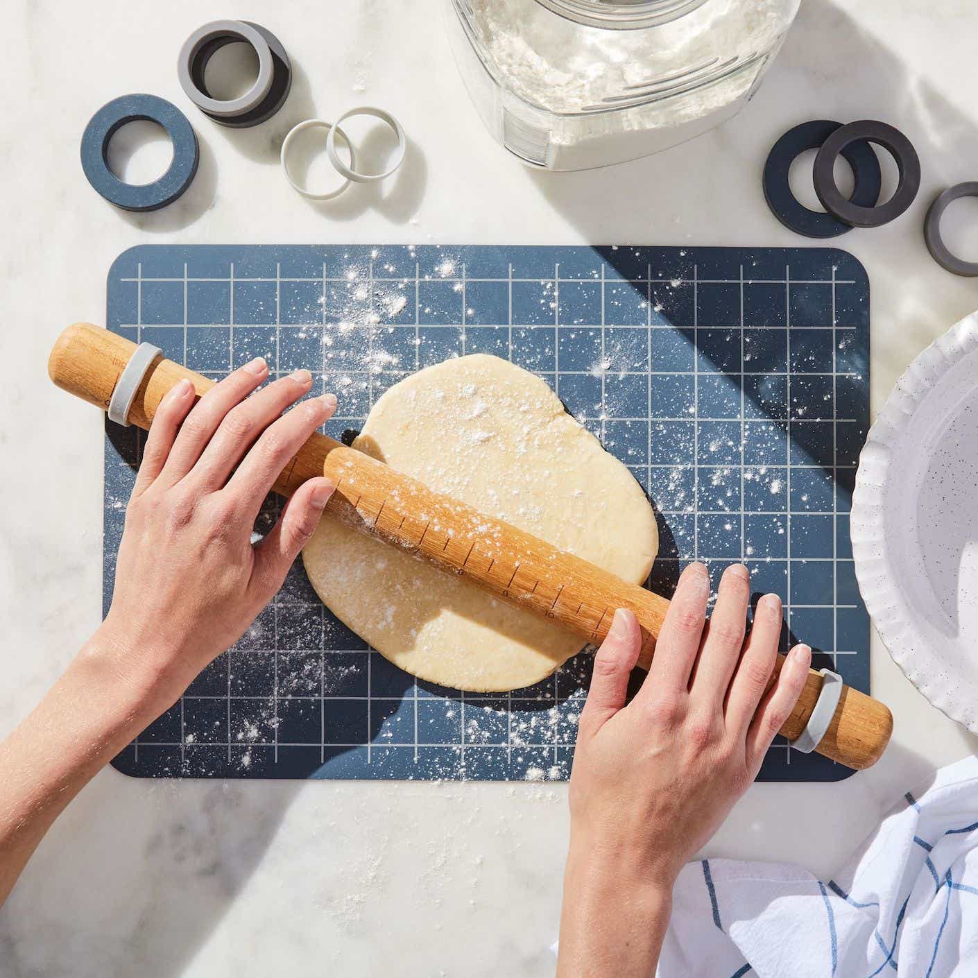 A pair of hands push a wooden rolling pin into a ball of dough that rests on a dark blue silicone baking mat.