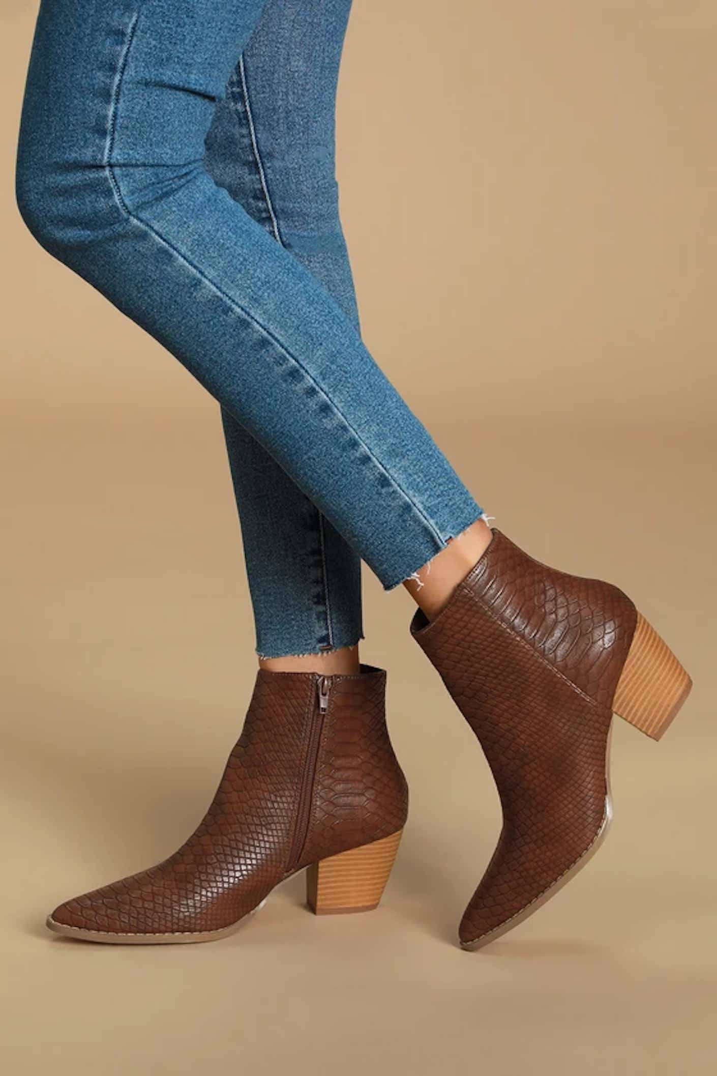 A person wearing jeans crosses their legs to show off dark brown snakeskin boots with a short, light brown heel.
