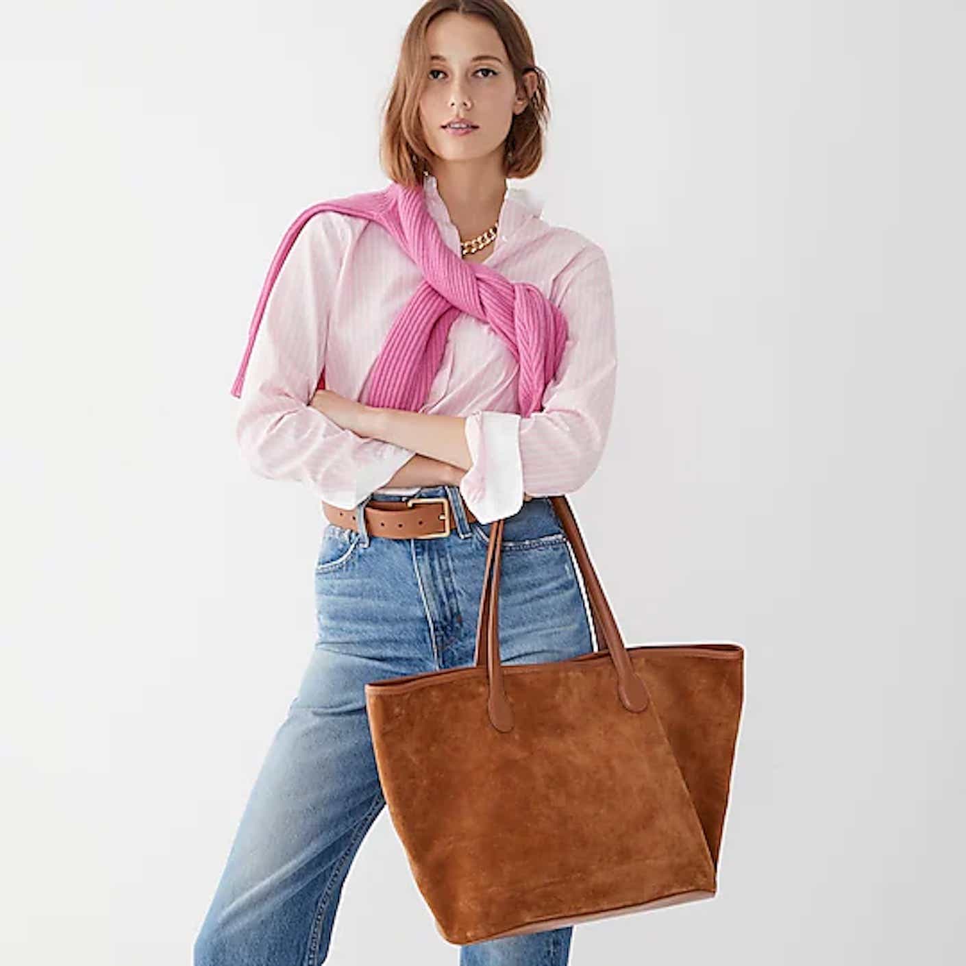 A woman wears a preppy outfit while holding a big, suede brown tote bag.
