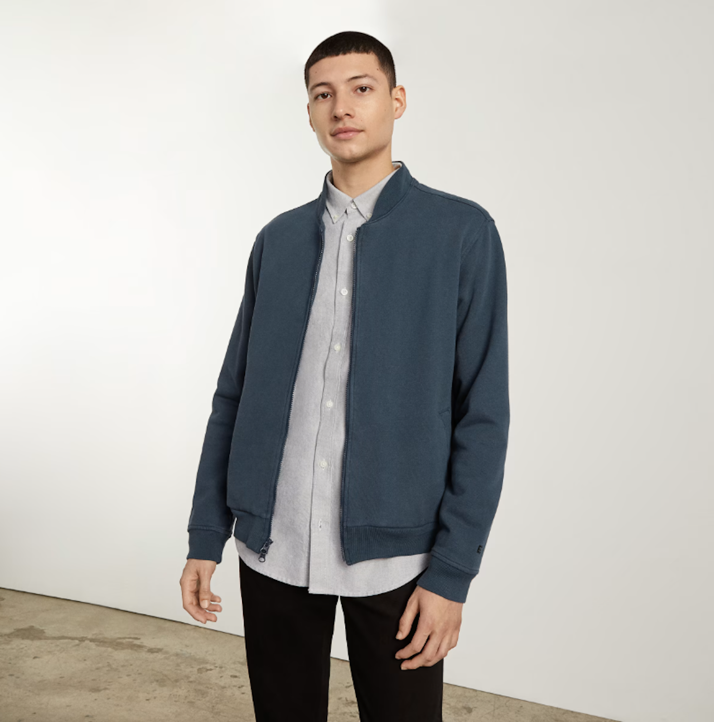 A man wears a classic, fitted slate blue track jacket over a button down shirt and jeans.