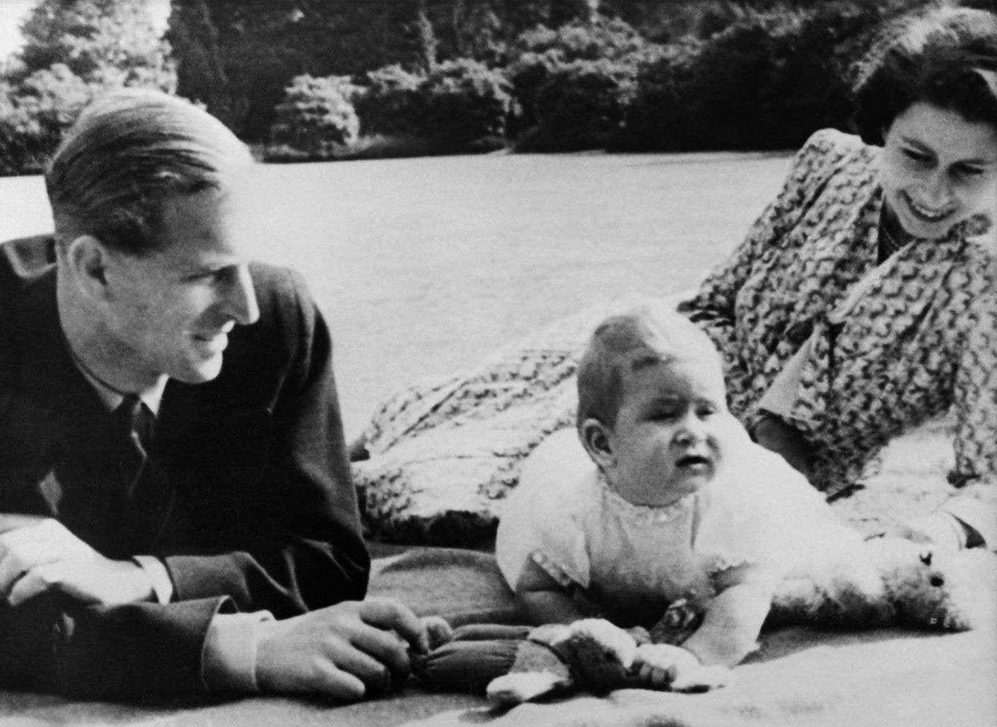 Princess Elizabeth of England and Prince Philip with their baby Prince Charles in 1949 lie togethr smiling on grass.