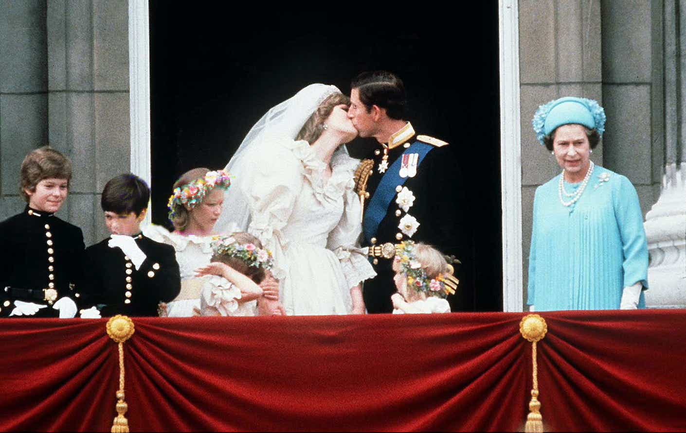 During their 1981 wedding, Prince Charles And Princess Diana kiss on the balcony of Buckingham Palace while Queen Elizabeth II stands to the side. Diana wears a long white wedding dress.