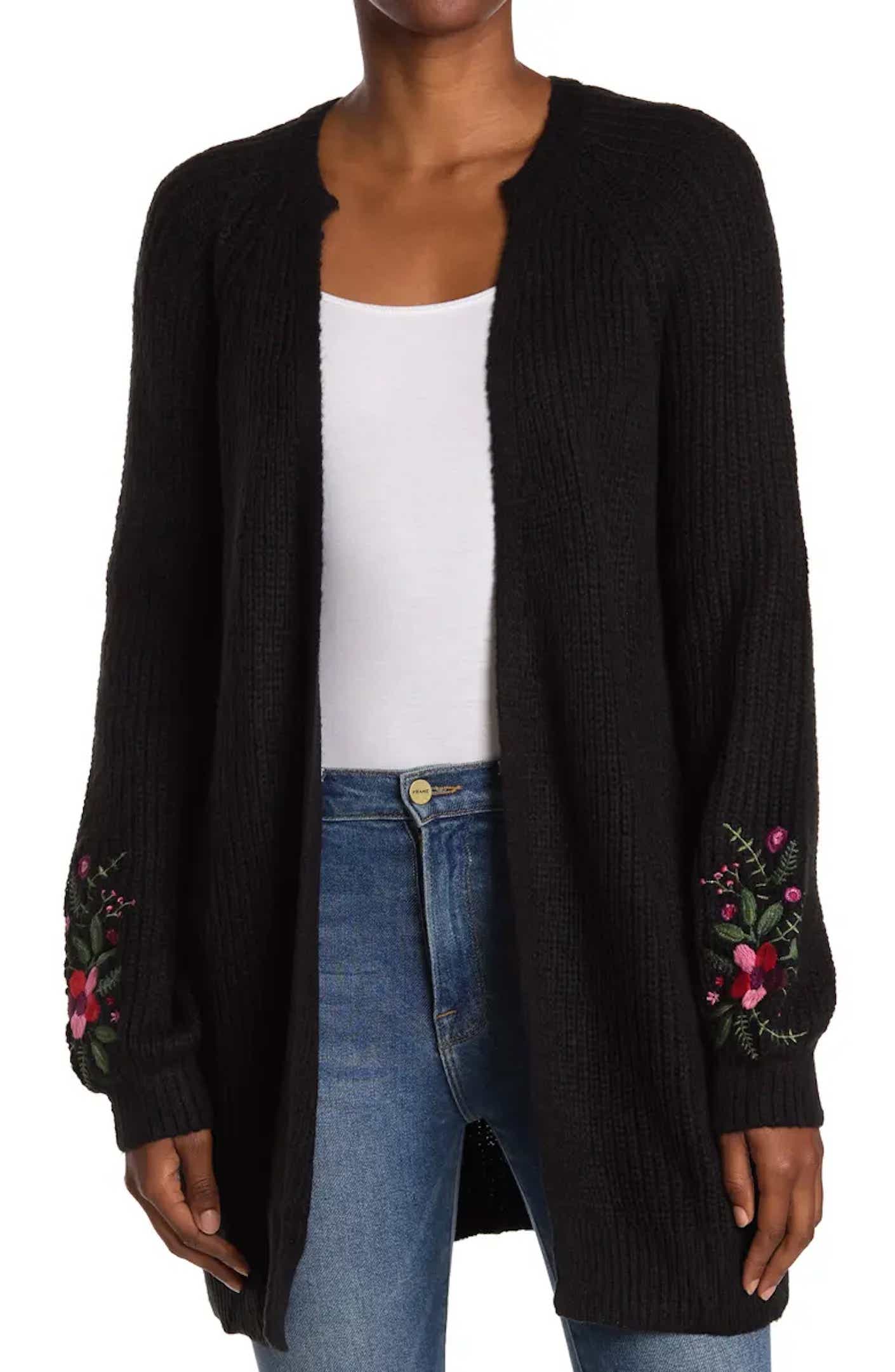 A woman wears an open front black cardigan with puffed long sleeves; the sleeves are embroidered with floral details.