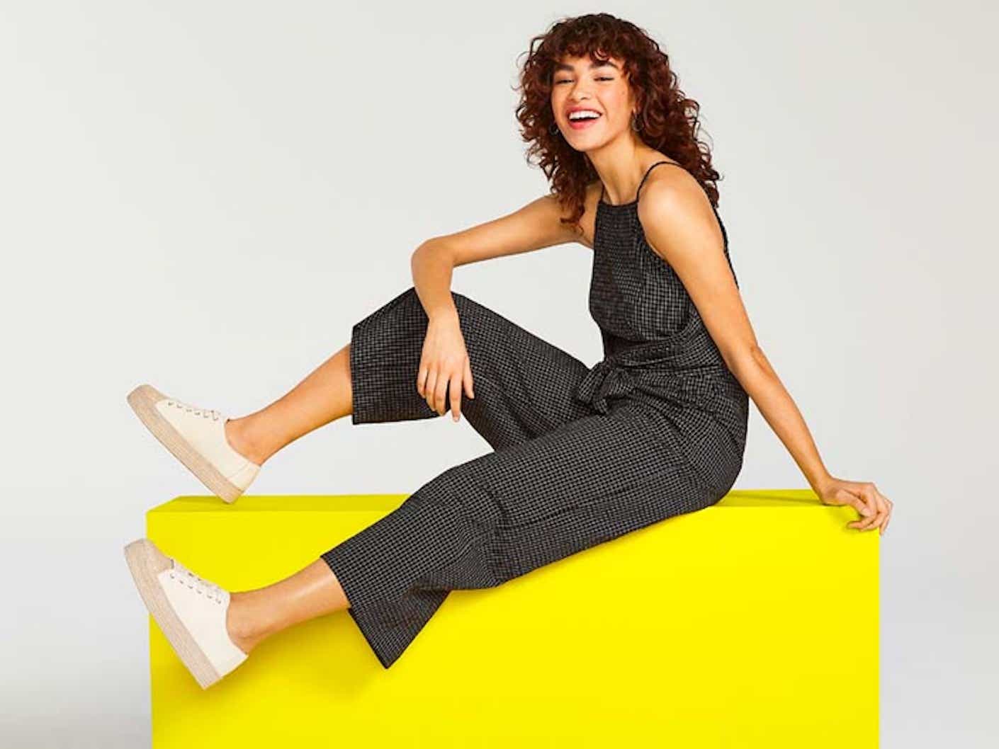 A laughing woman sits on a yellow block while wearing a black jumpsuit with white sneakers.
