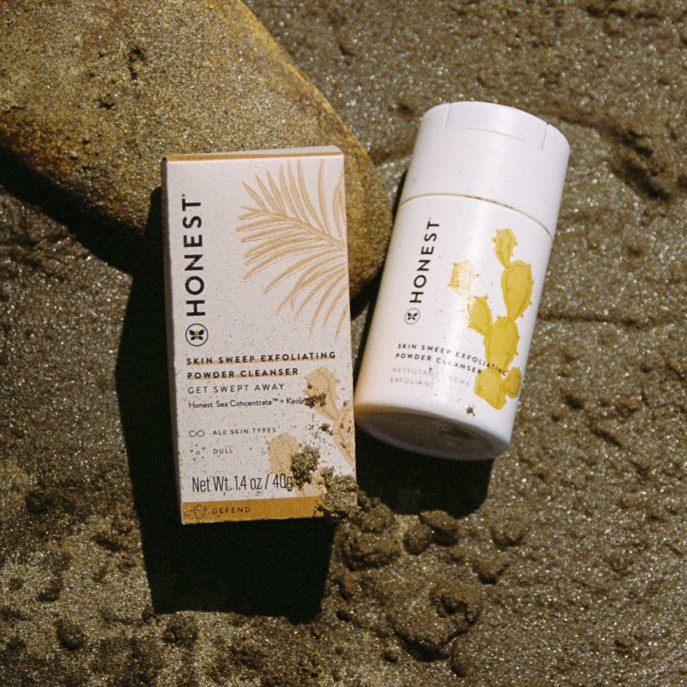 A cylindrical container of powder cleanser lies in a sunny patch of sand.