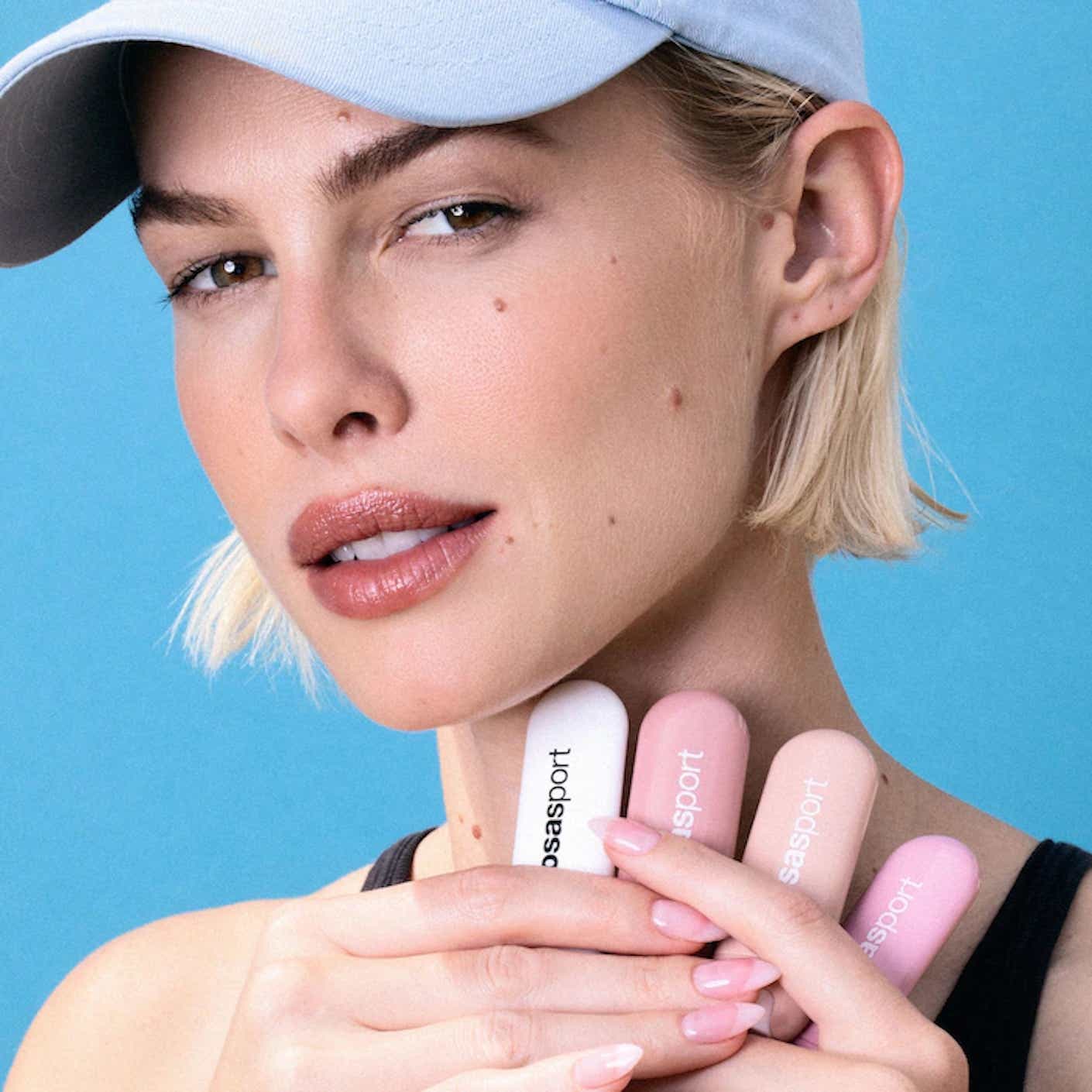 A woman holds four tubes of lip balm up to her lightly tinted and unchapped lips.