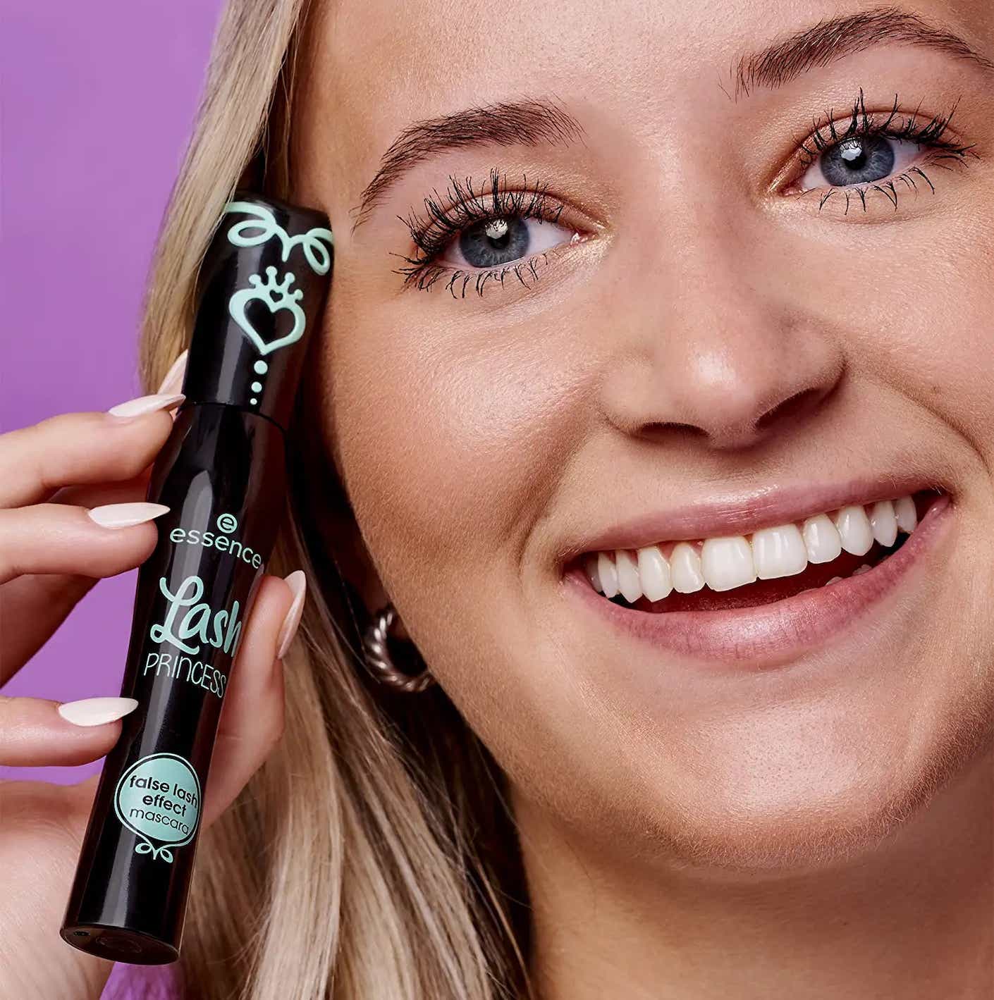 A smiling woman with long, black eyelashes holds a black tube of mascara up next to her face.