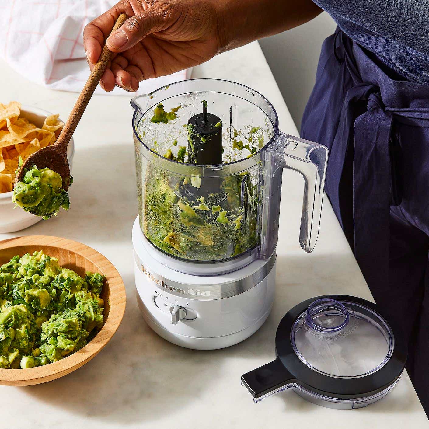 A small, cordless food processor sits on a countertop while filled with guacamole that a person spoons out of the food processor.
