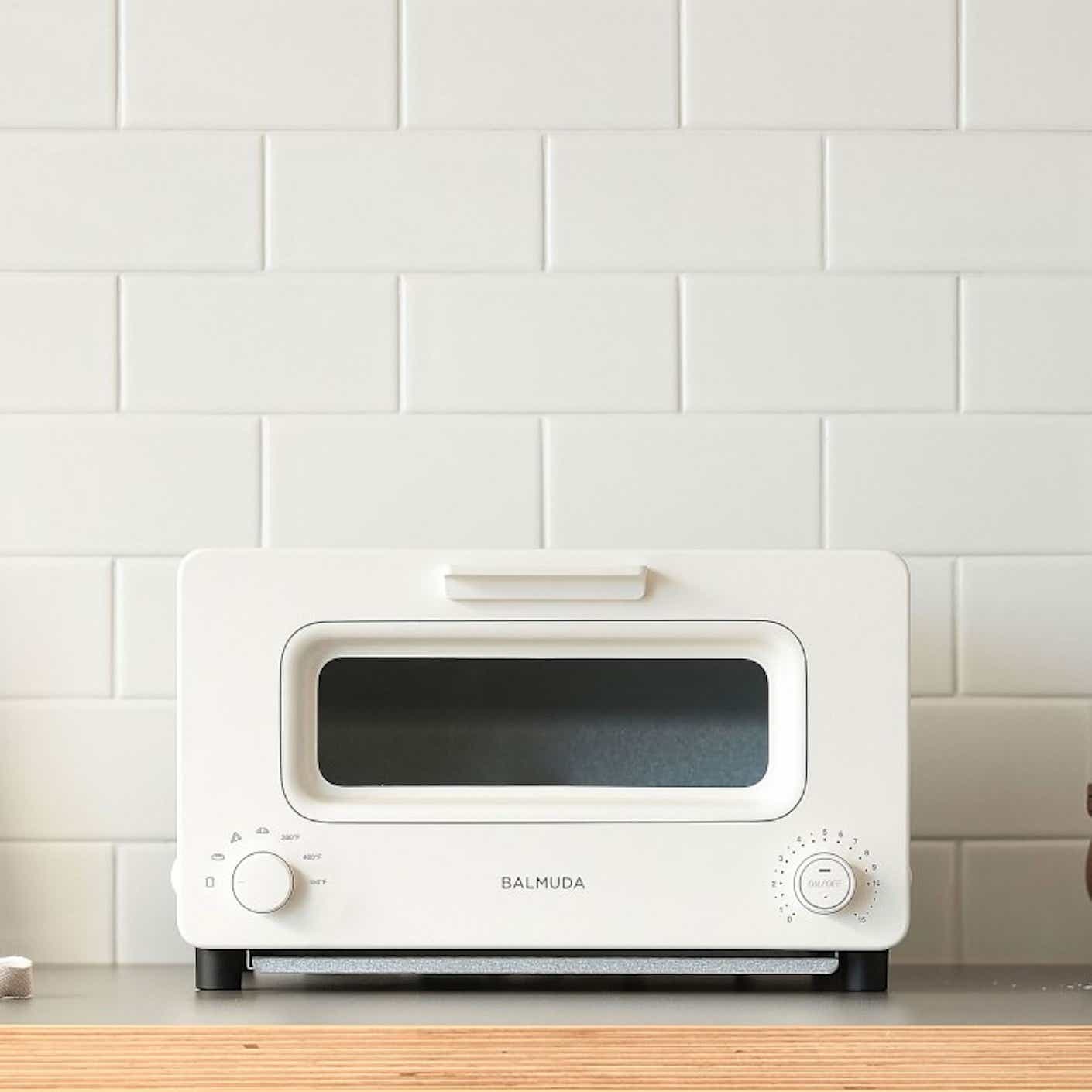 A white toaster oven with knobs sits on a plain countertop in front of a white tiled wall.