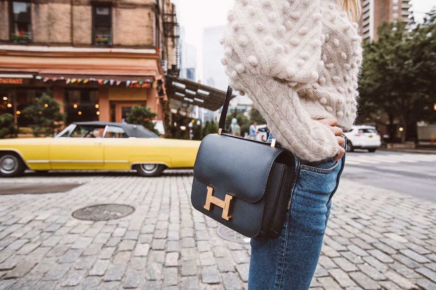 A woman wears a black handbag with gold details in front of a cobblestone lined street.