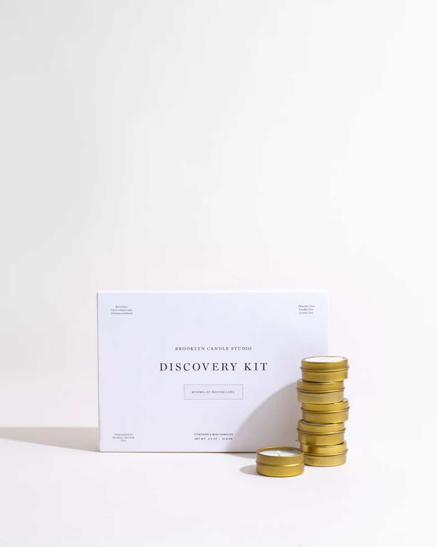 A stack of white, scented tea candles in bronze colored metallic containers sits in front of a minimalistic box.