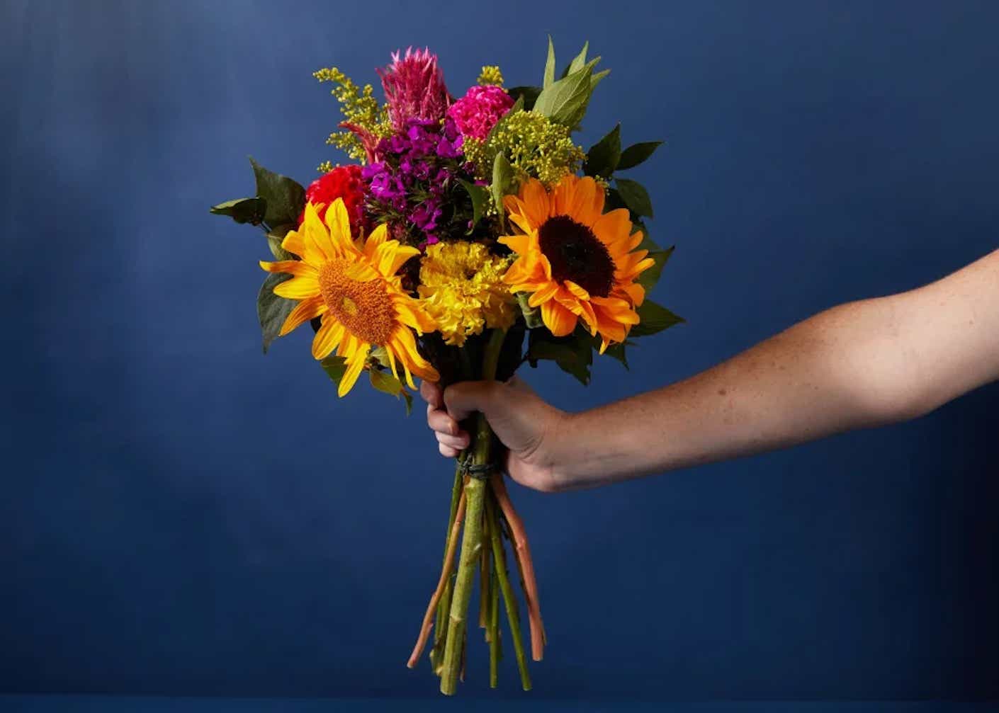 A disembodied arm holds a bouquet of mixed, bright flowers with different colors and textures.