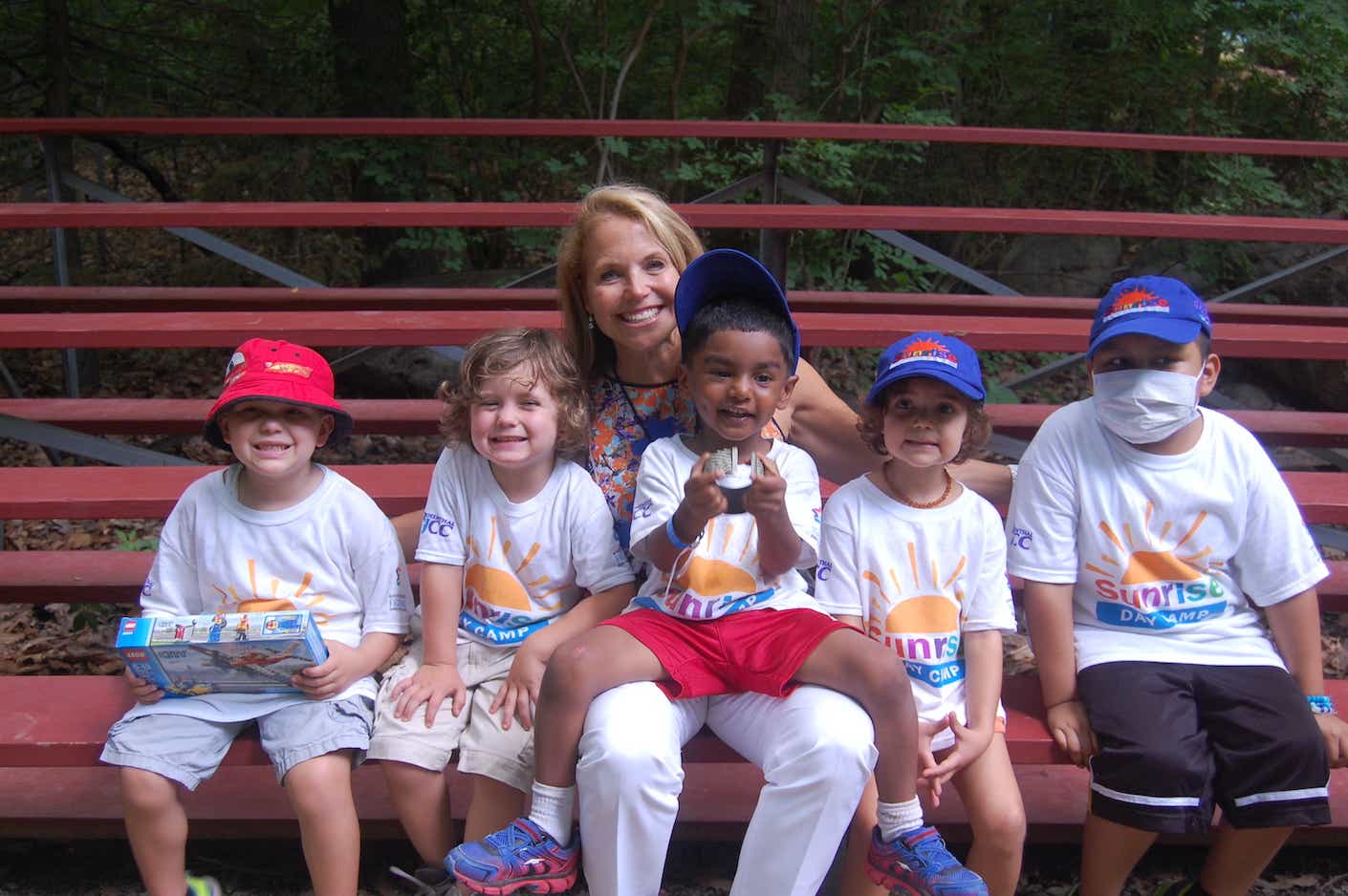 Katie Couric smiles and holds several young campers on a set of red bleachers.