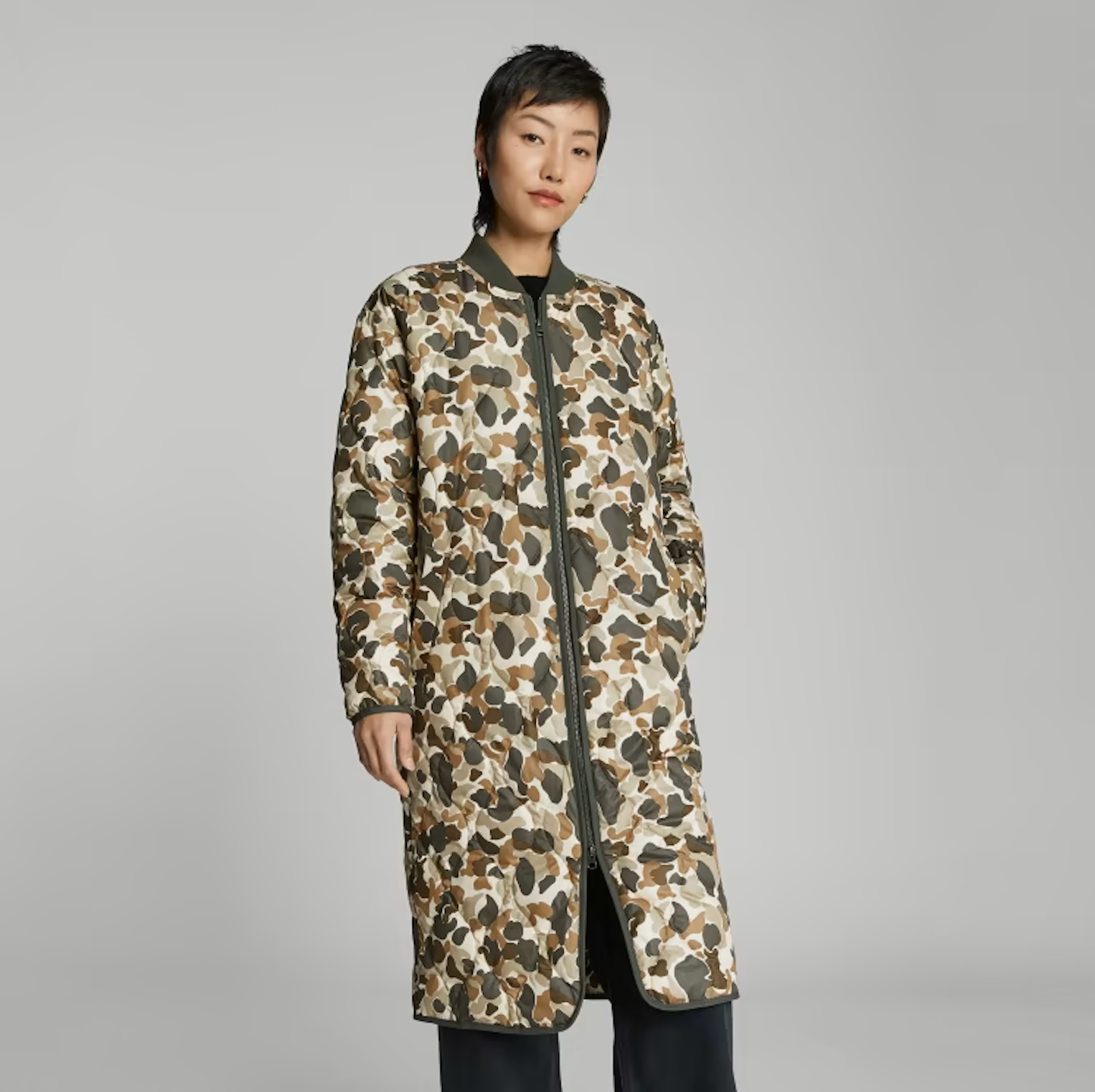 A woman wears a long, midweight, quilted coat with blobby, green, white, and brown camo print.