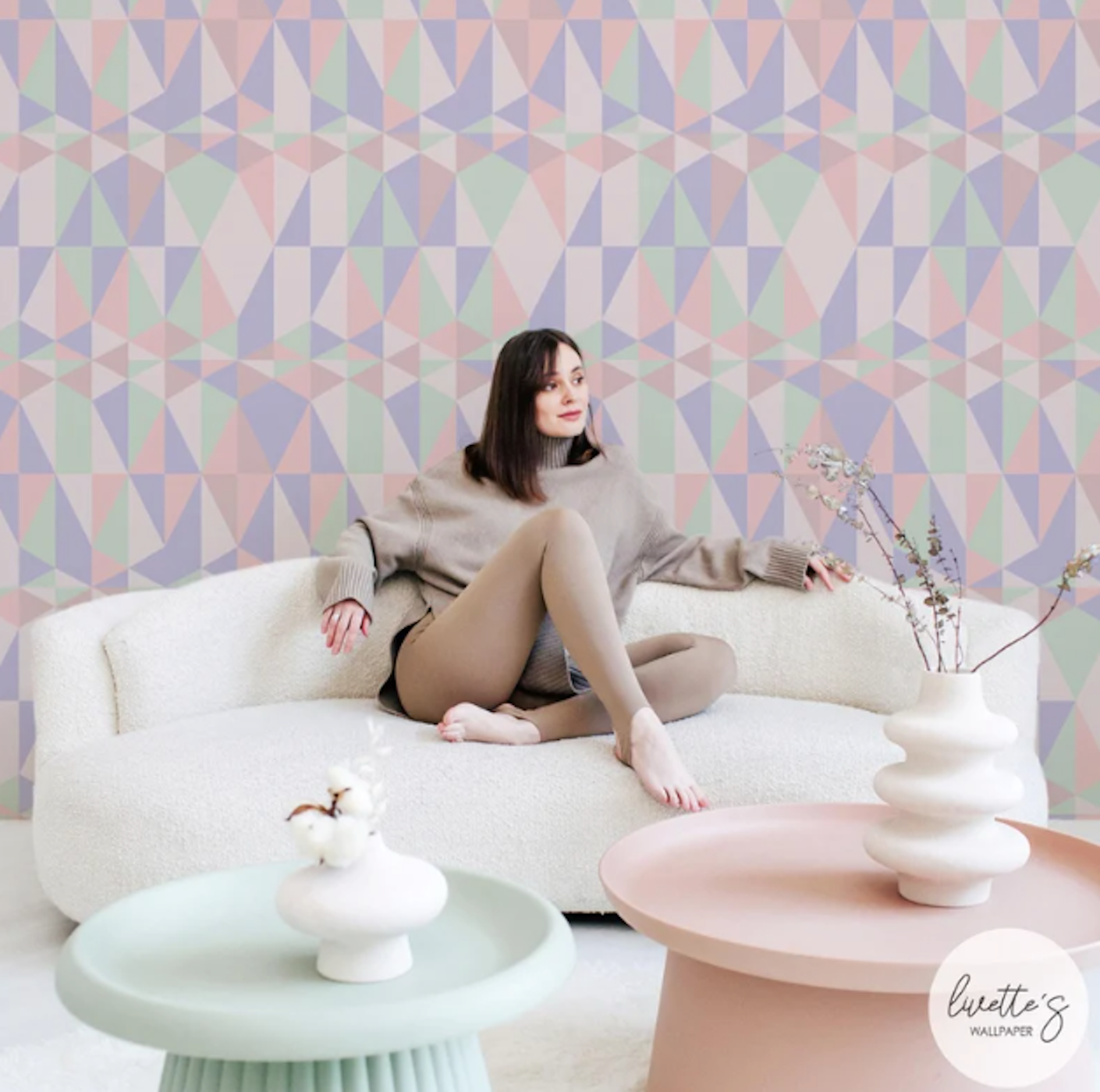 A woman lounges in a living space filled with pastel furniture in front of a swatch of pastel geometric wallpaper.