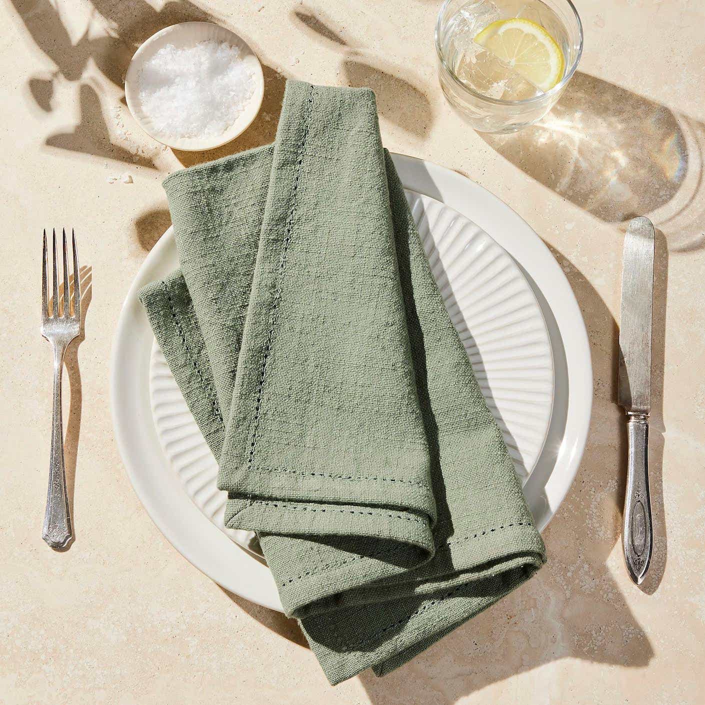 A sage green napkin is folded neatly on a white plate that sits on a cream table cloth.