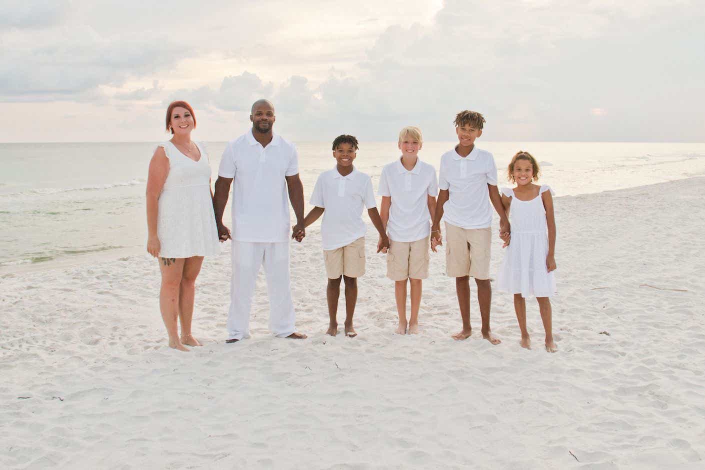 A family of six stands smiling on a white sand beach at dusk, holding hands and wearing matching white outfits.