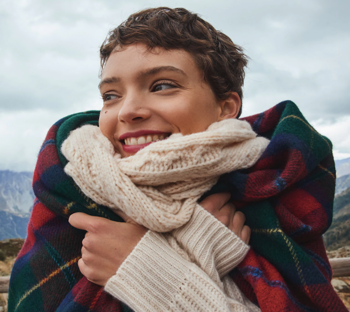 A smiling woman huddles up in a blanket and scarf.