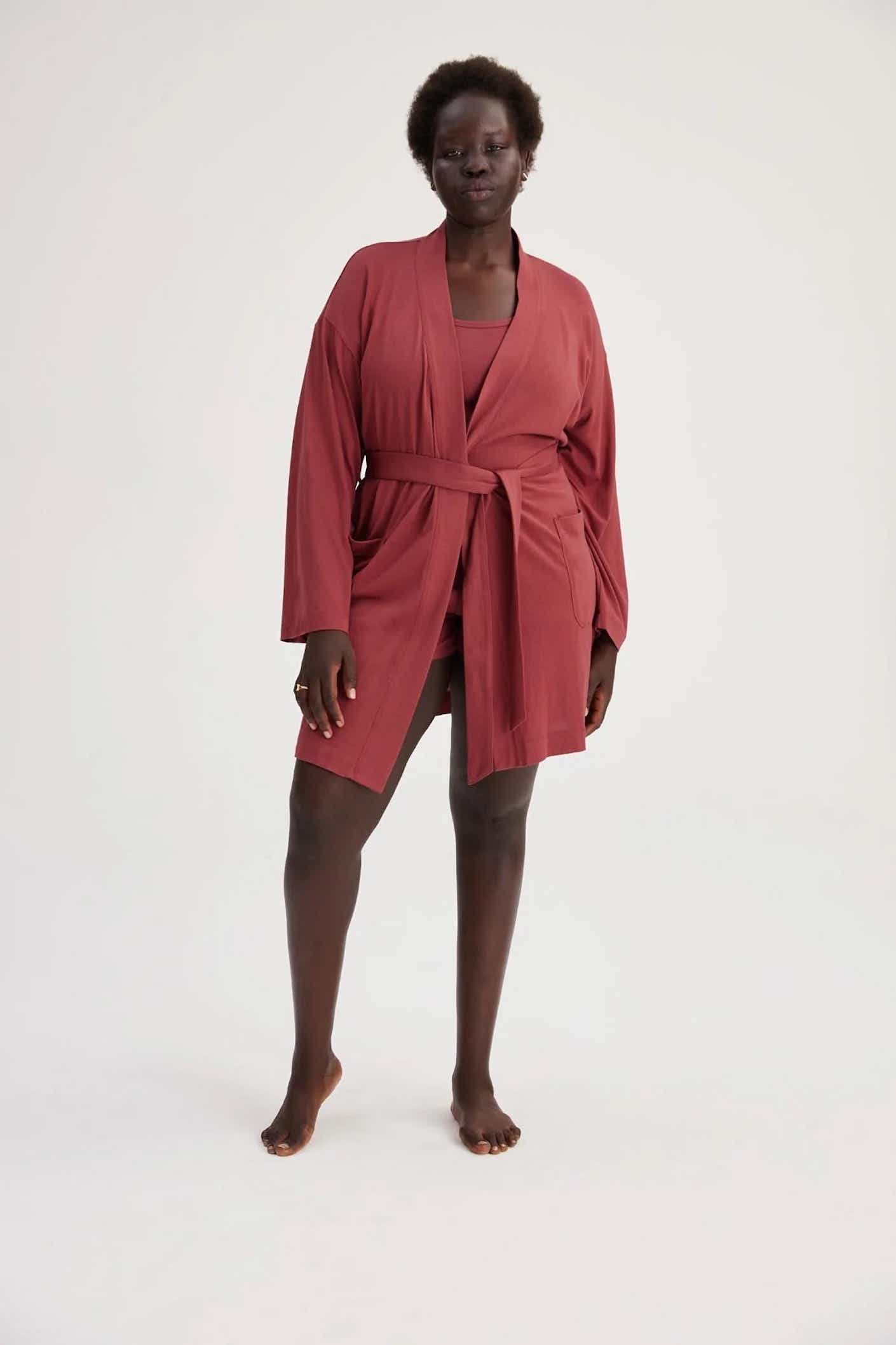 A woman wears a deep red robe that hits at mid thigh while standing in front of a grey background.