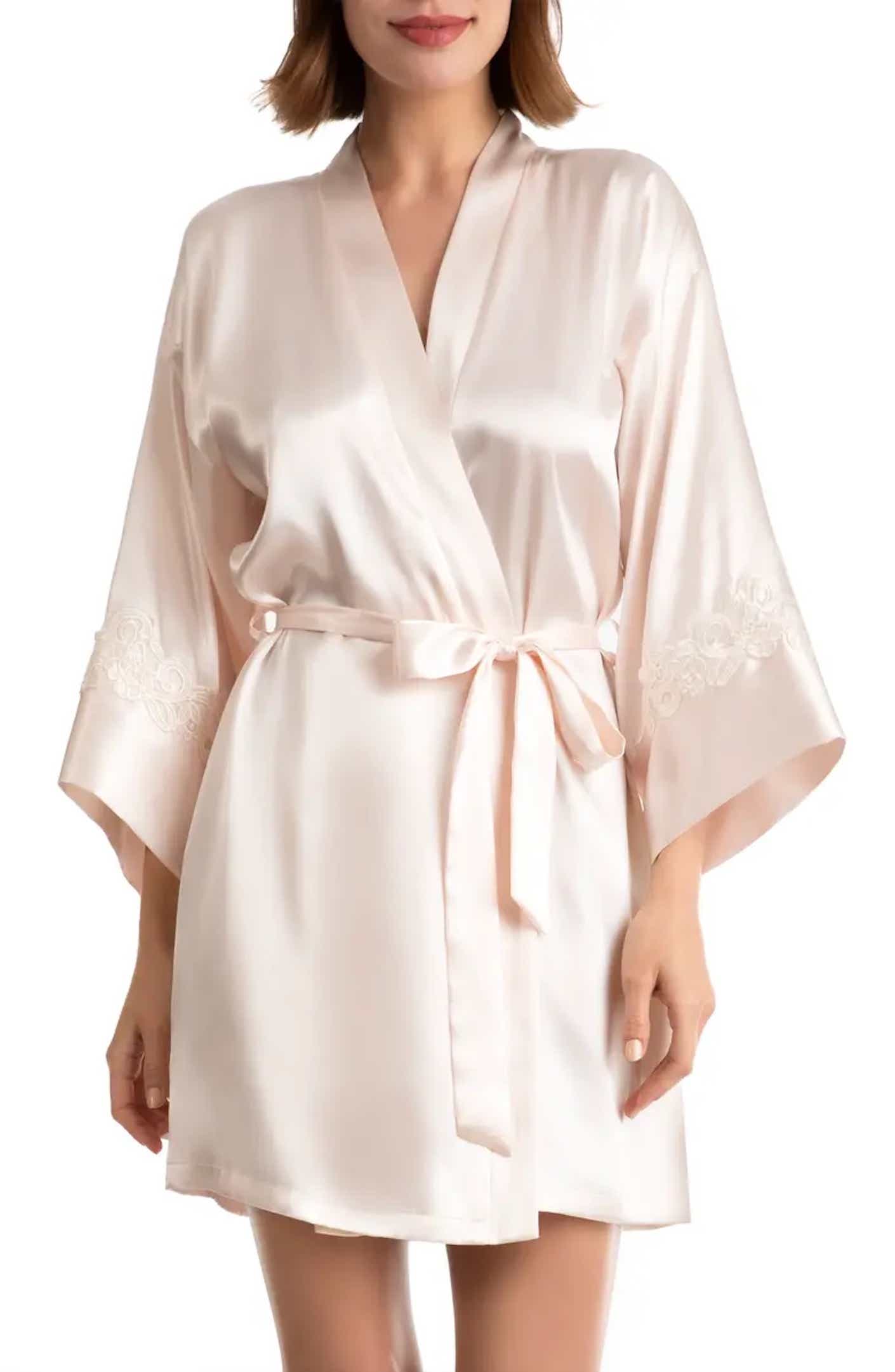 A woman wears a thigh length satin robe in a light peach color in front of a white background.
