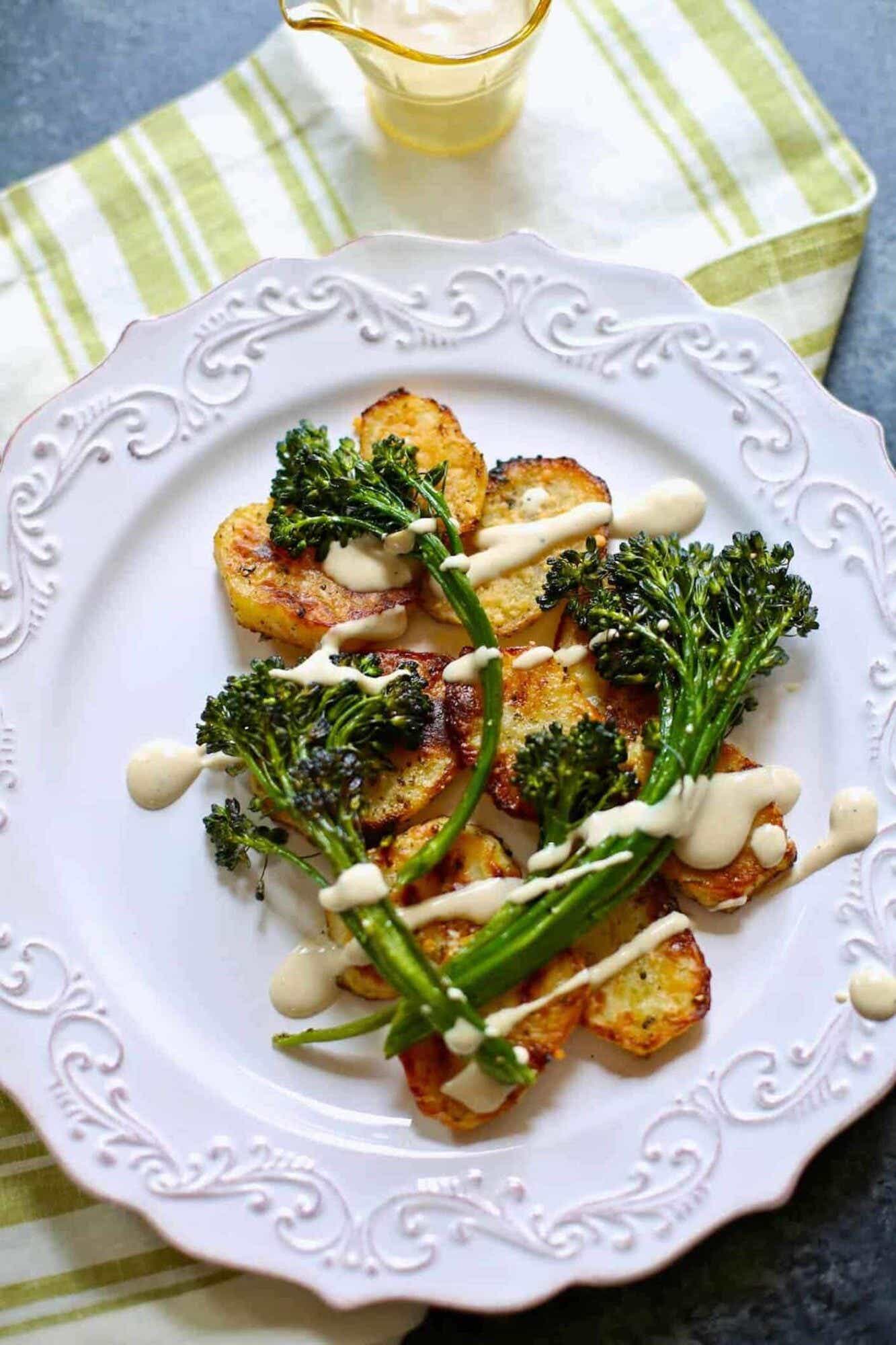 A white china plate is full of roasted potatoes and broccolini and sprinkled with a white sauce.