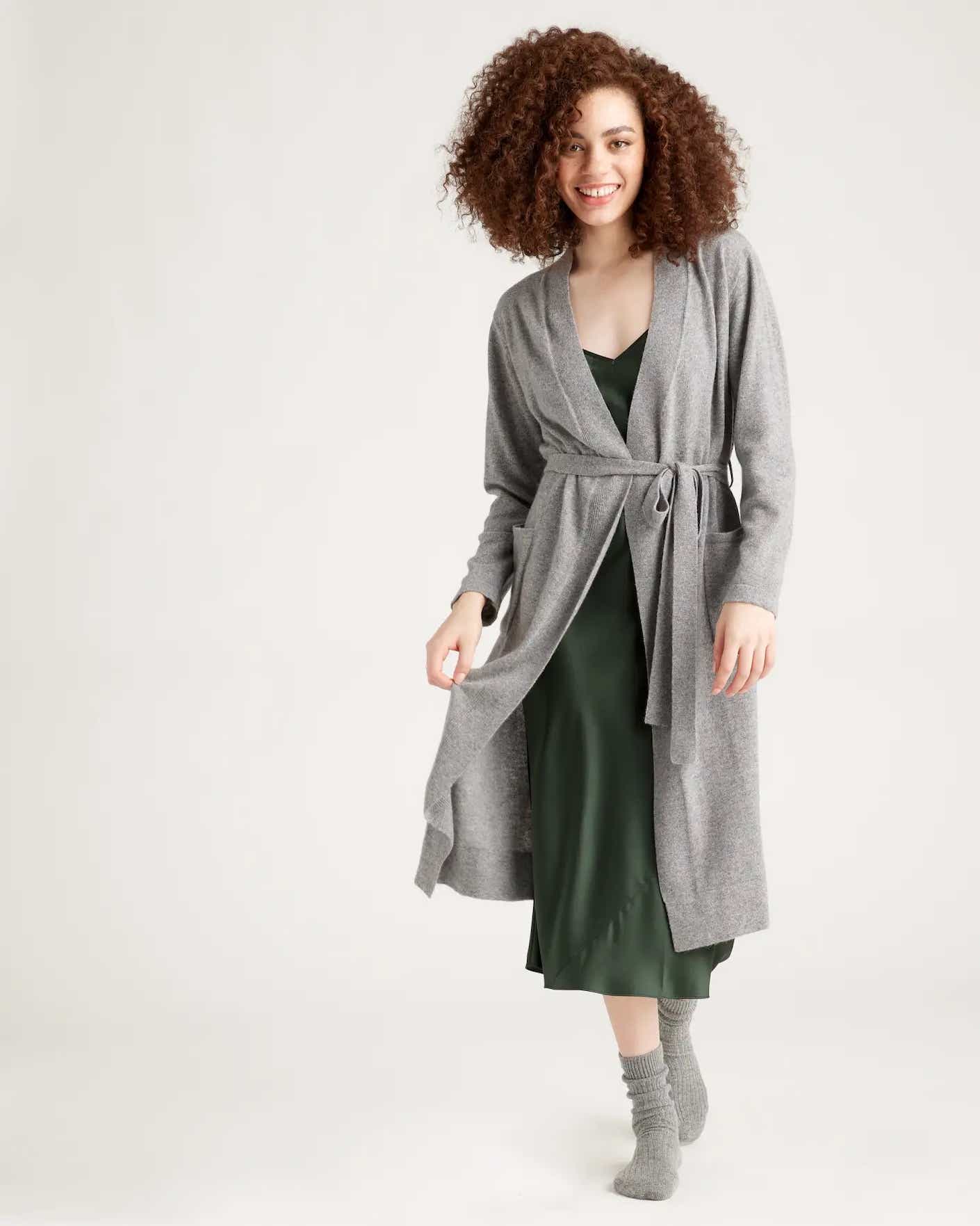A woman smiles as she steps forward in a light grey, mid weight, cashmere robe that falls to her shins.