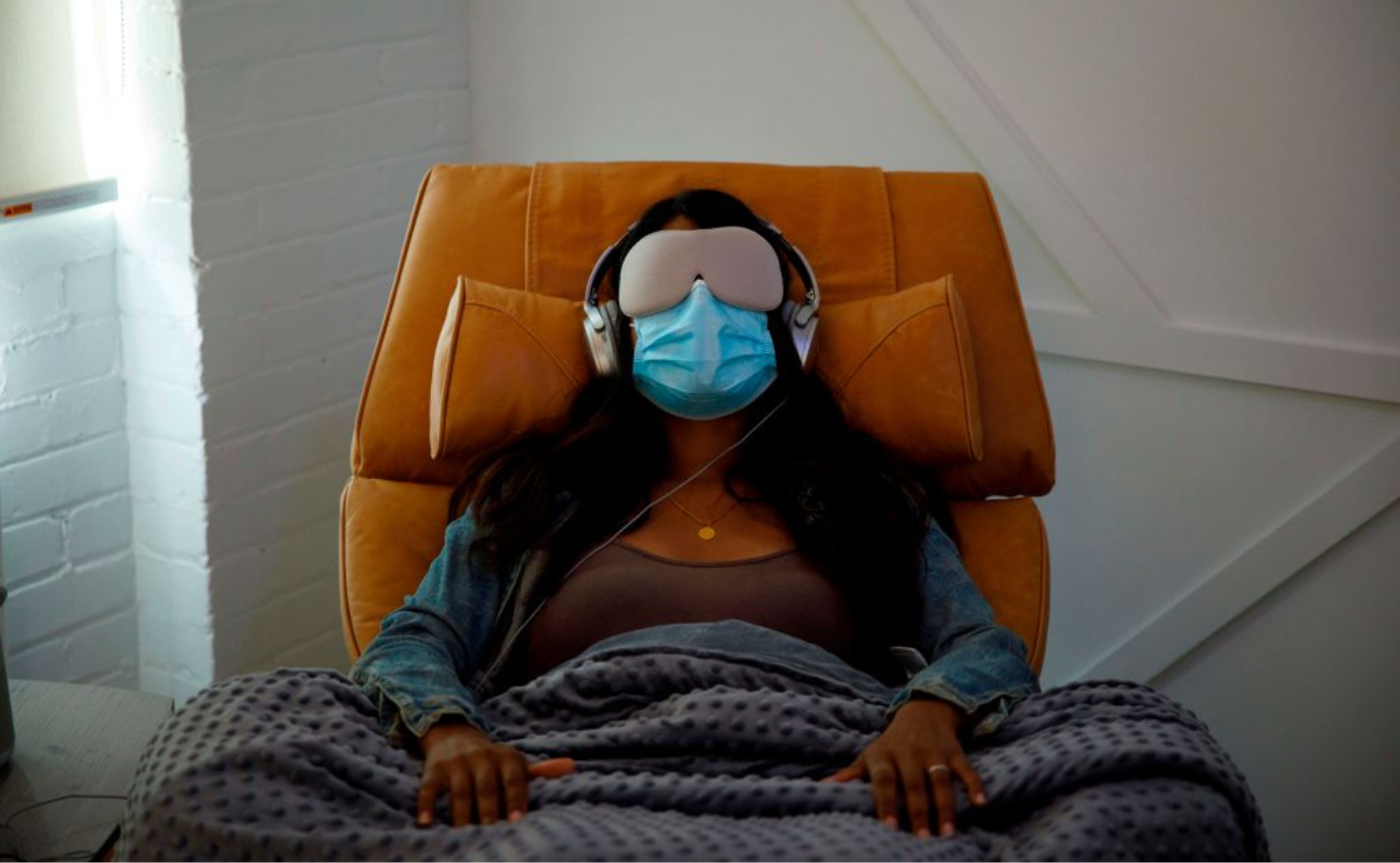 A woman in a chair with a blanket, glasses, and headphones, at a psychedelic medicine clinic
