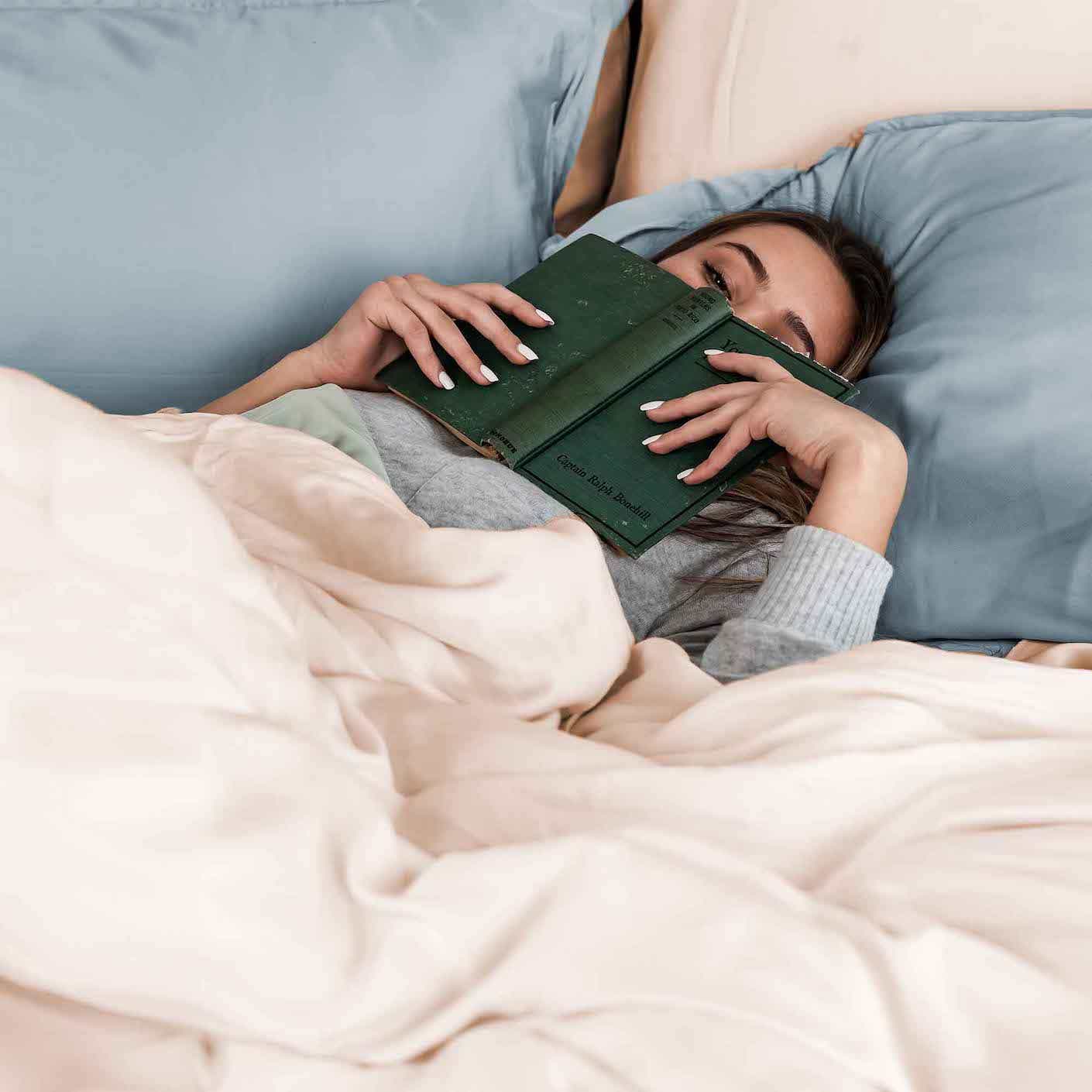 A woman in soft luxurious sheets relaxes with a book over her face.