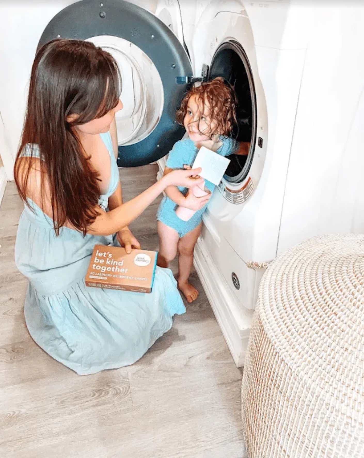 A woman and child kneel in front of a dryer with laundry sheets in hand.