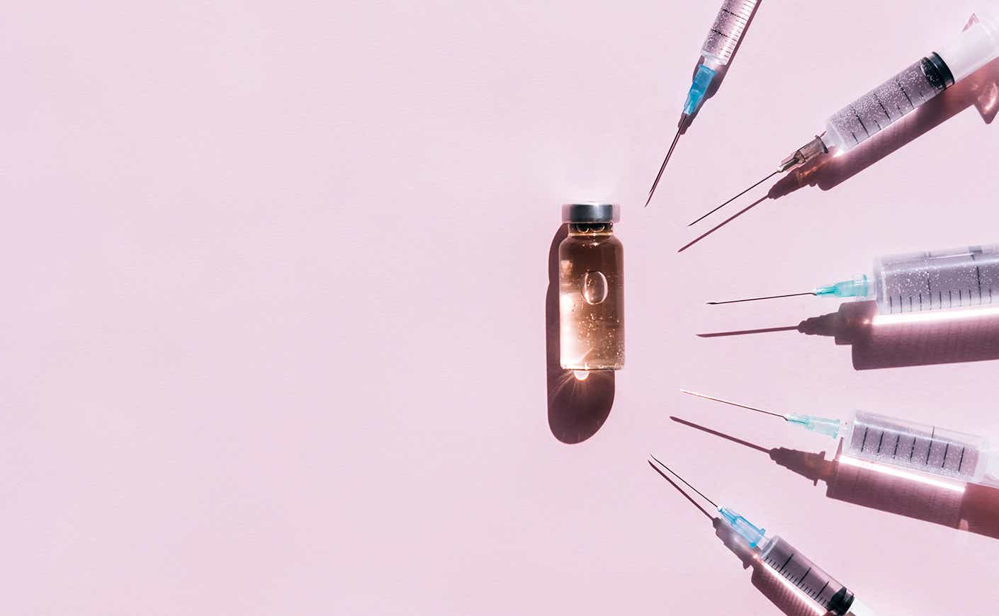 four syringes facing in towards vial of clear liquid on pink background