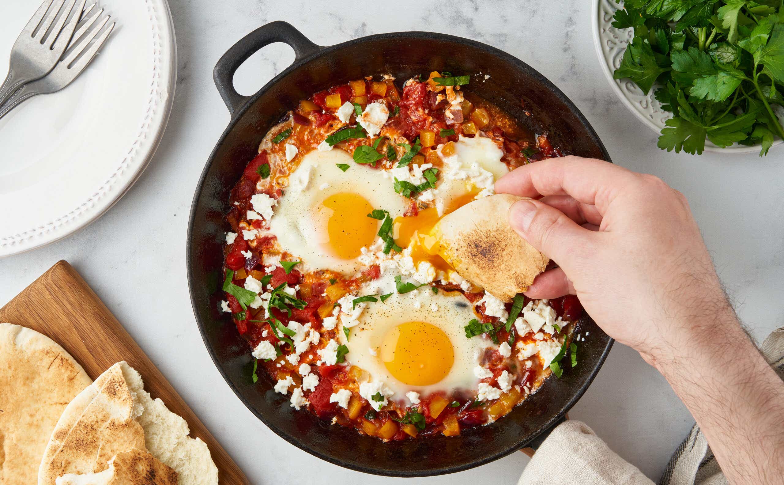 Male hand dipping flatbread into shakshouka in frying pan.